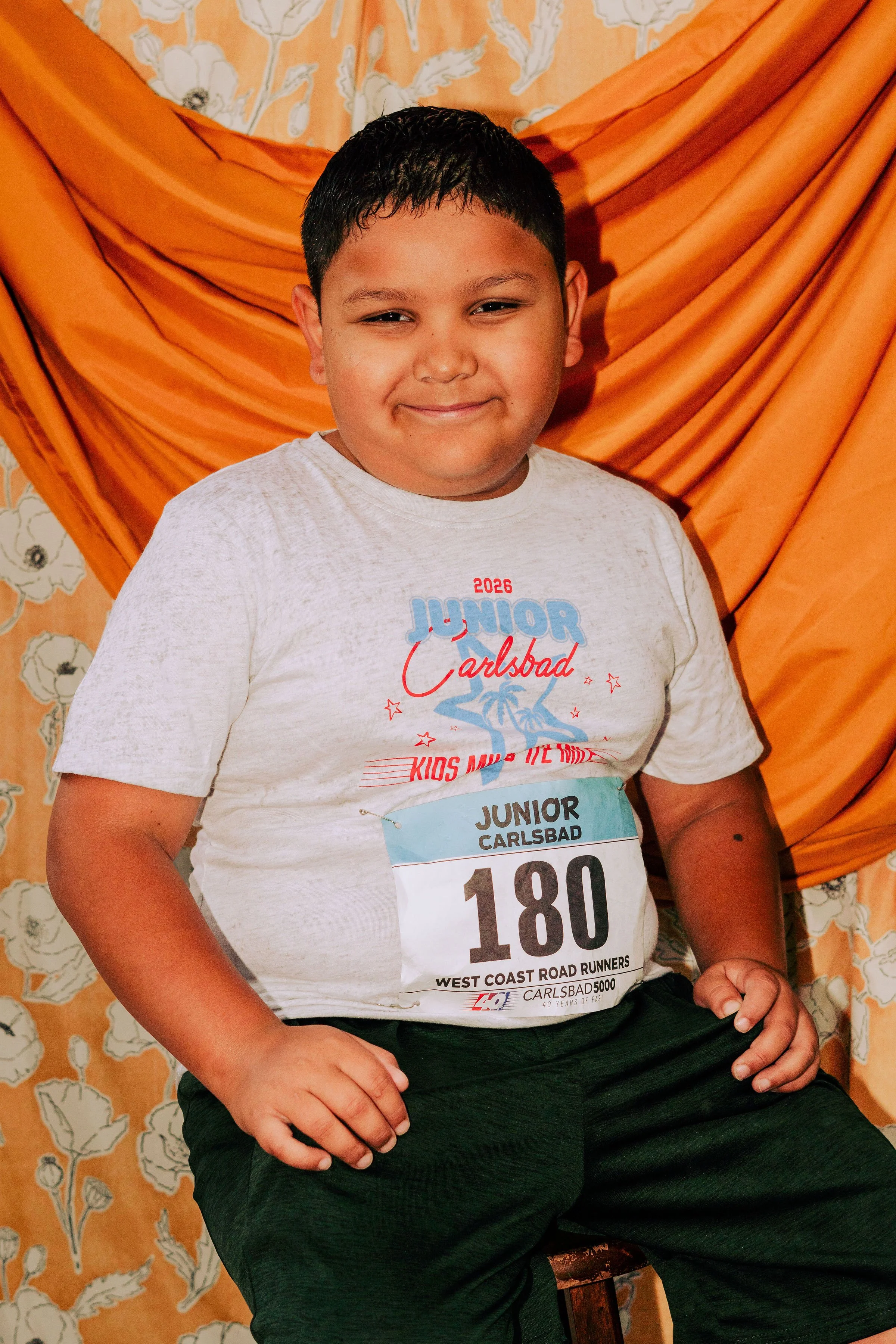 A young boy smiling, wearing a white t-shirt with race number 180, sitting in front of orange drapes and floral wallpaper.