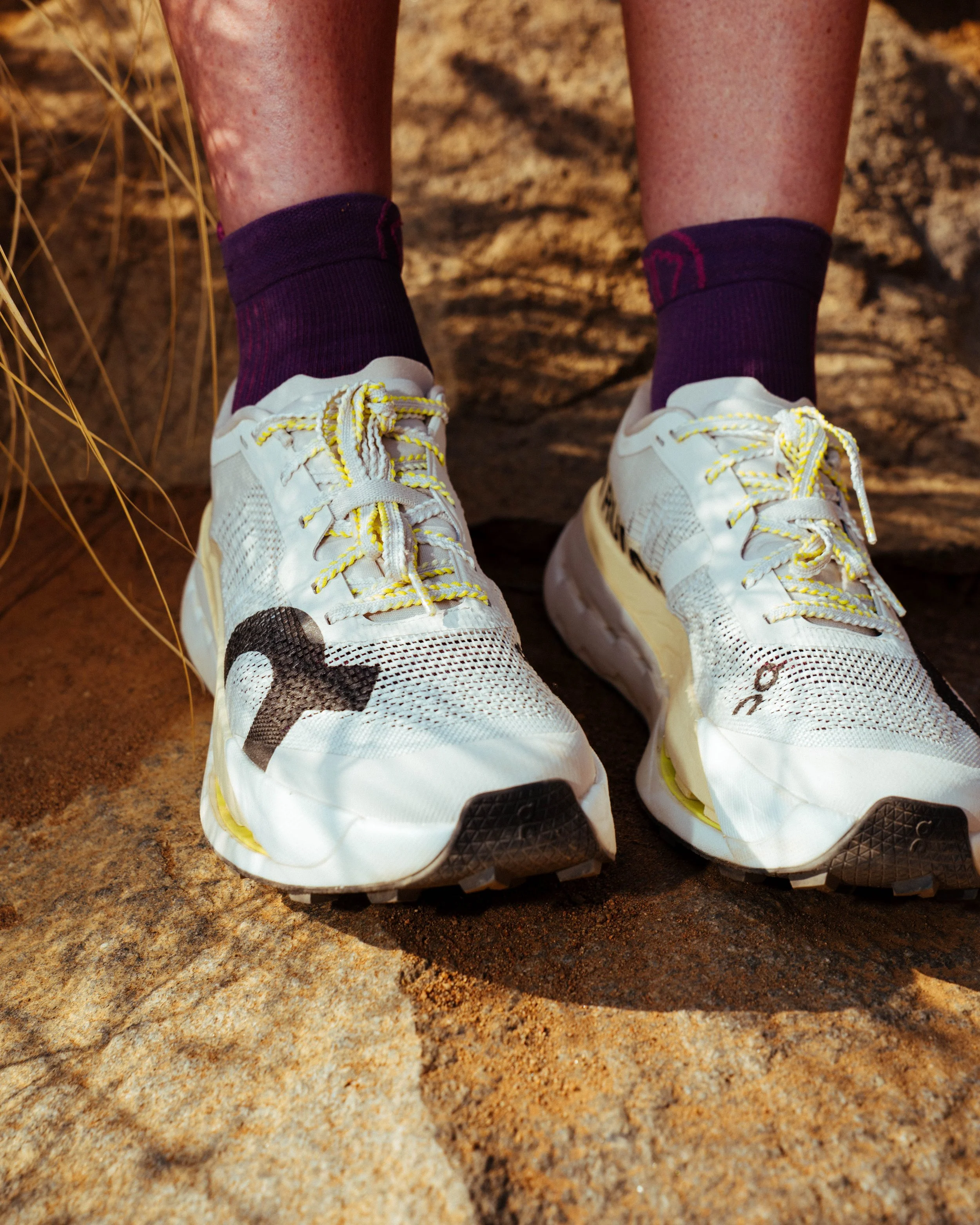 Close-up of a person's feet wearing white running shoes with yellow laces, purple socks, and sitting on a rocky surface.