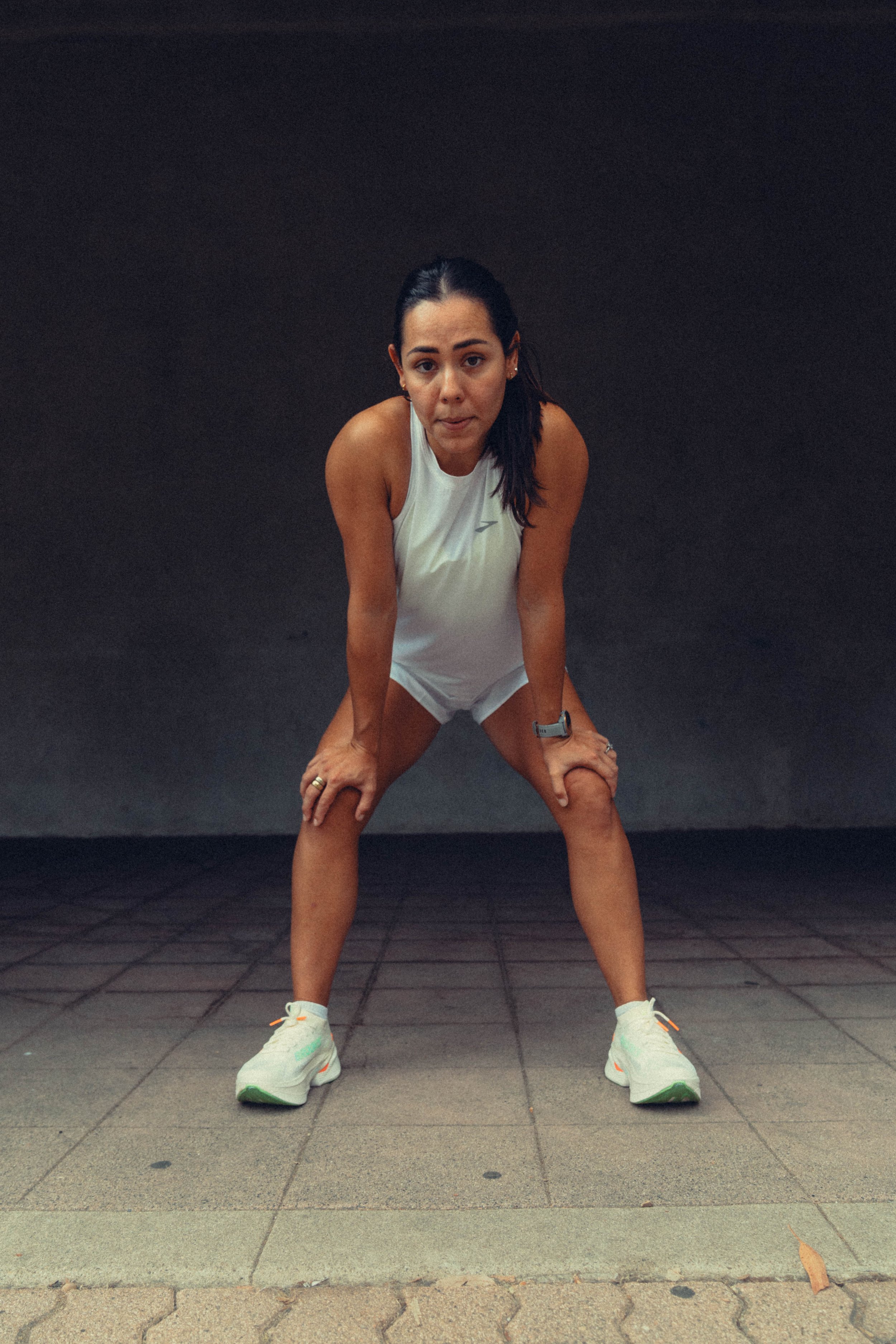 Woman in white tank top and shorts stretching in front of black wall, wearing athletic shoes and a watch.