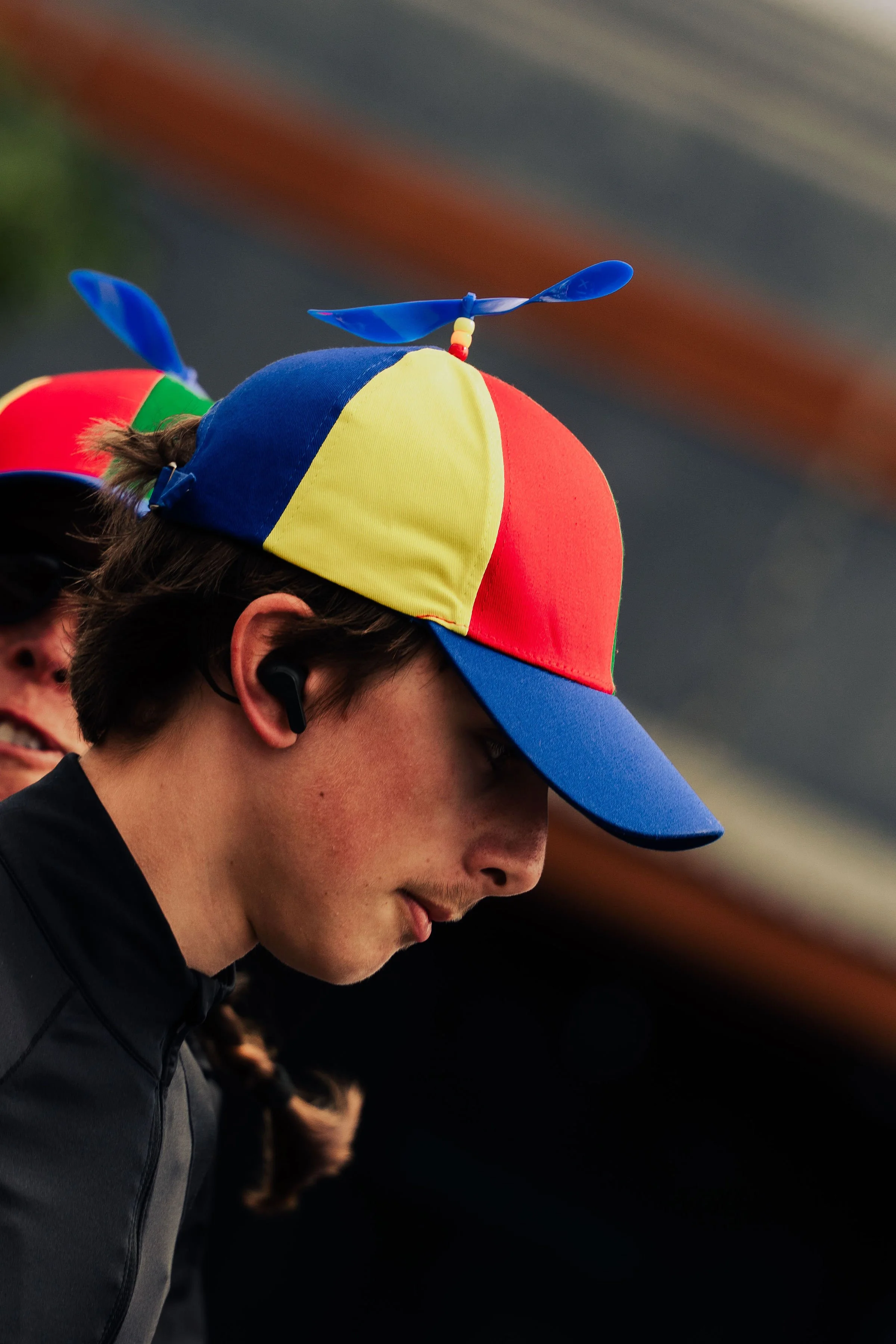 Close-up side view of a young man wearing a multicolored cap with a small blue propeller on top, black jacket, and wireless earbud, in a dimly lit environment.