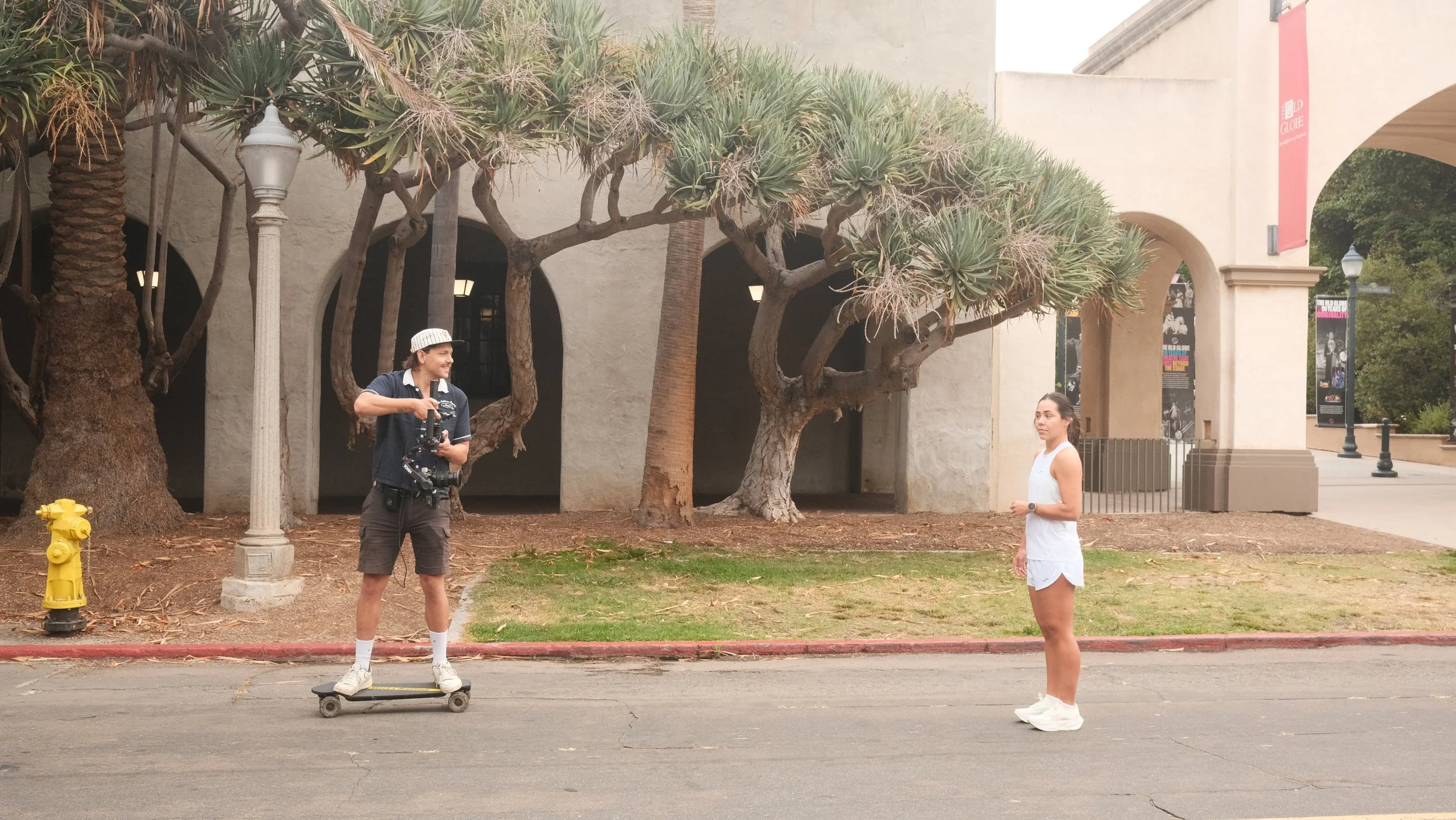 A man on a skateboard taking a selfie with a young woman in a white athletic outfit outdoors near a row of trees and a building.