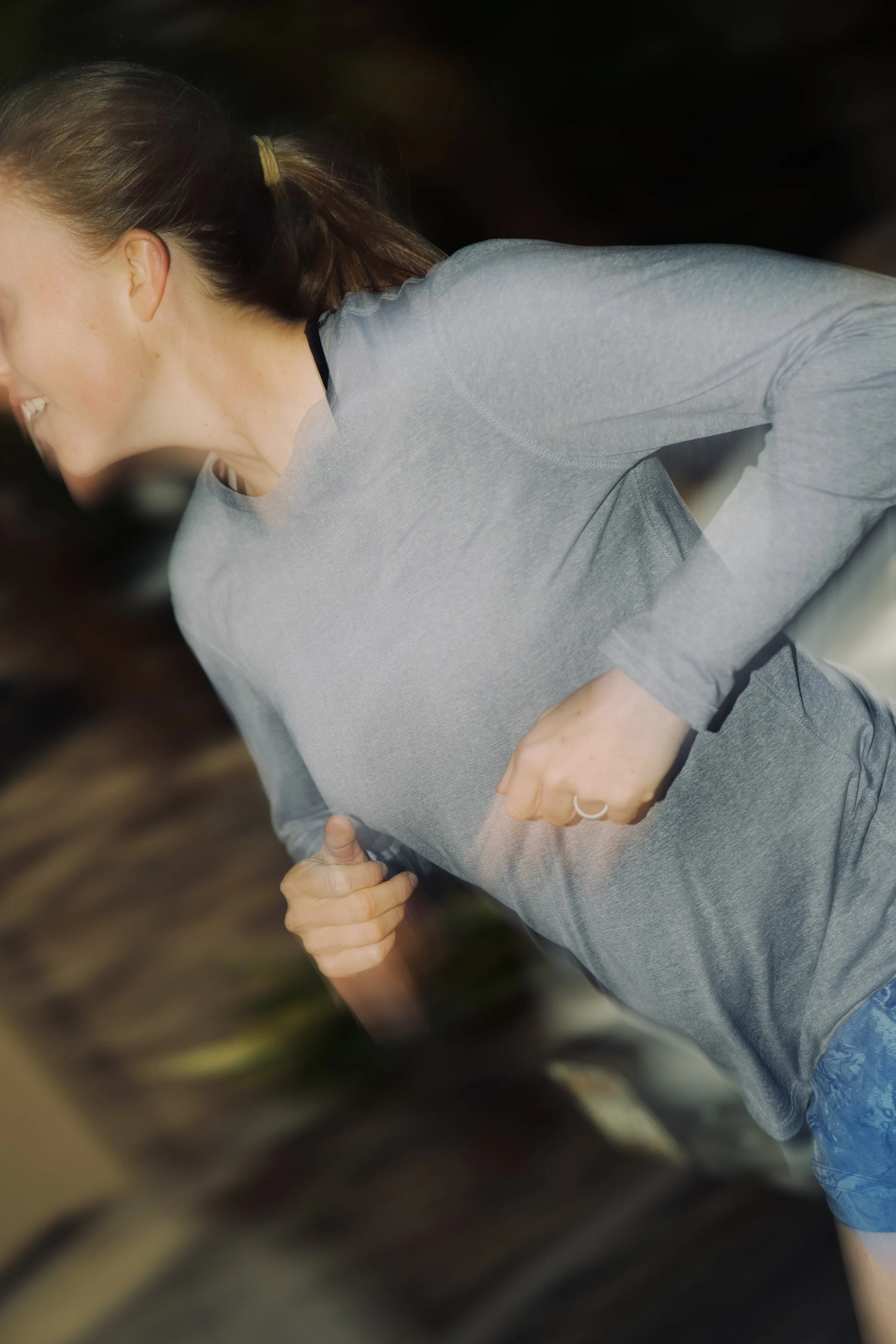 A woman with brown hair tied in a ponytail, wearing a gray long-sleeve shirt, smiling and looking to her right while holding her waist with her left hand, in an indoor setting.