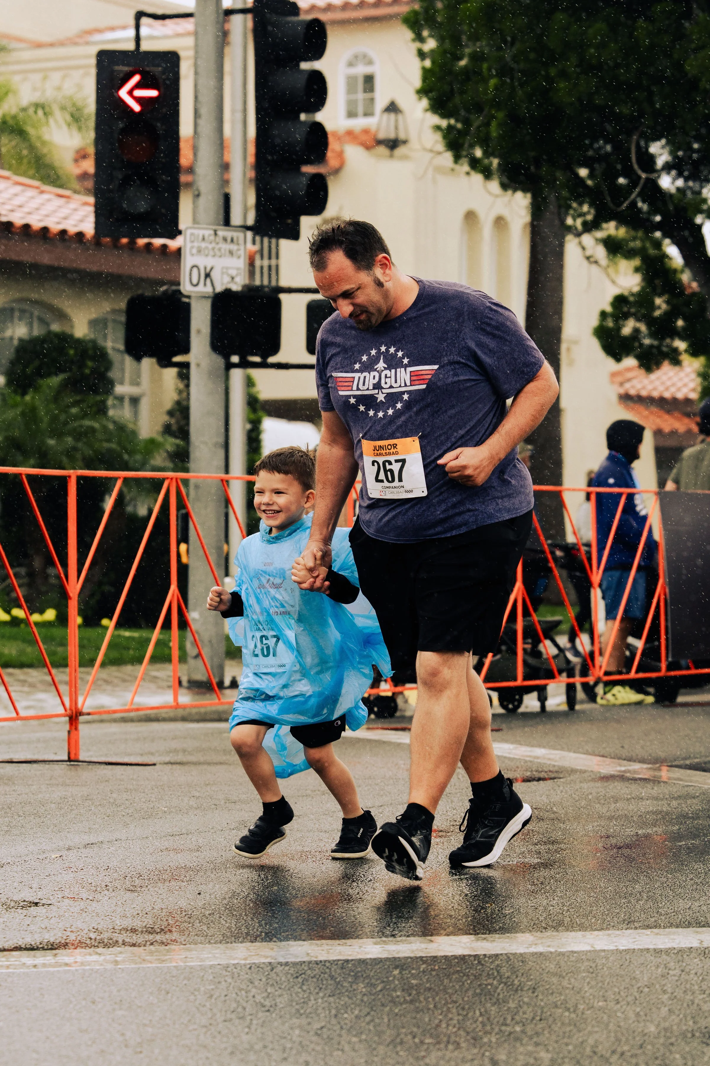 A man and a young boy smiling and holding hands while running in a street race during rainy weather, with traffic lights and buildings in the background.