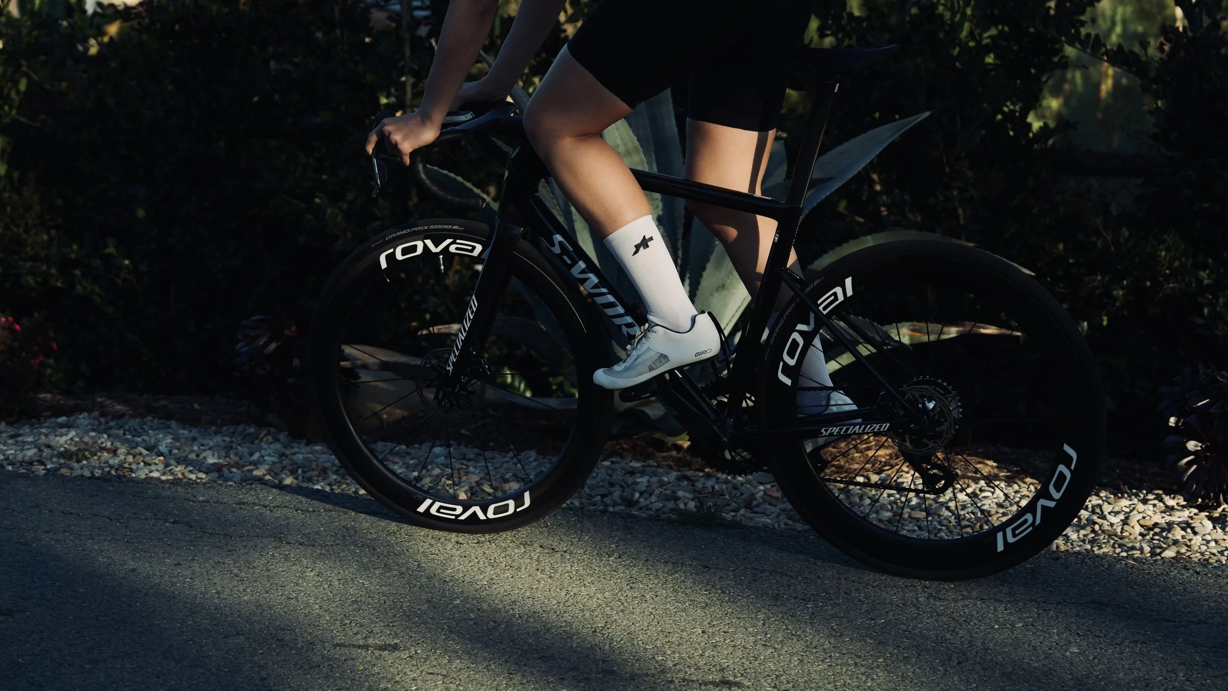 Close-up of a cyclist riding a black Specialized S-Works Tarmac road bike, wearing white socks with a black logo and white shoes, on a gravel path with greenery in the background.