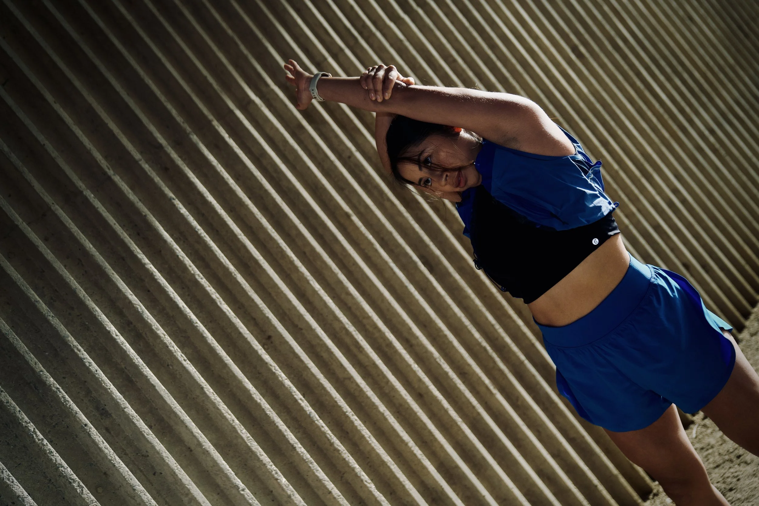 A woman in athletic clothing stretching her arm above her head against a background of concrete stairs.