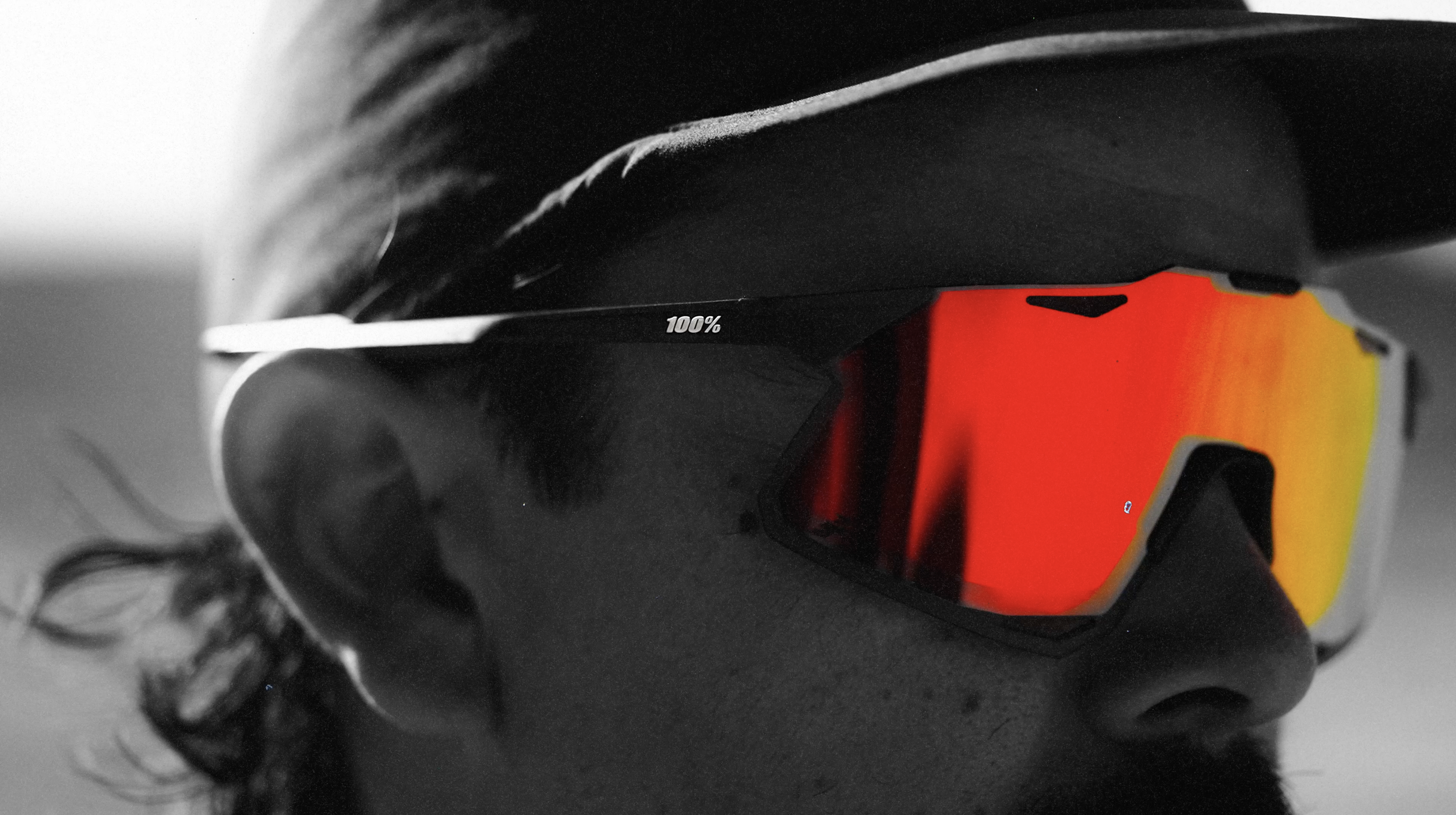 A pair of black and red ski goggles with a 100% label on the frame, resting on a dark surface next to curling black hair.