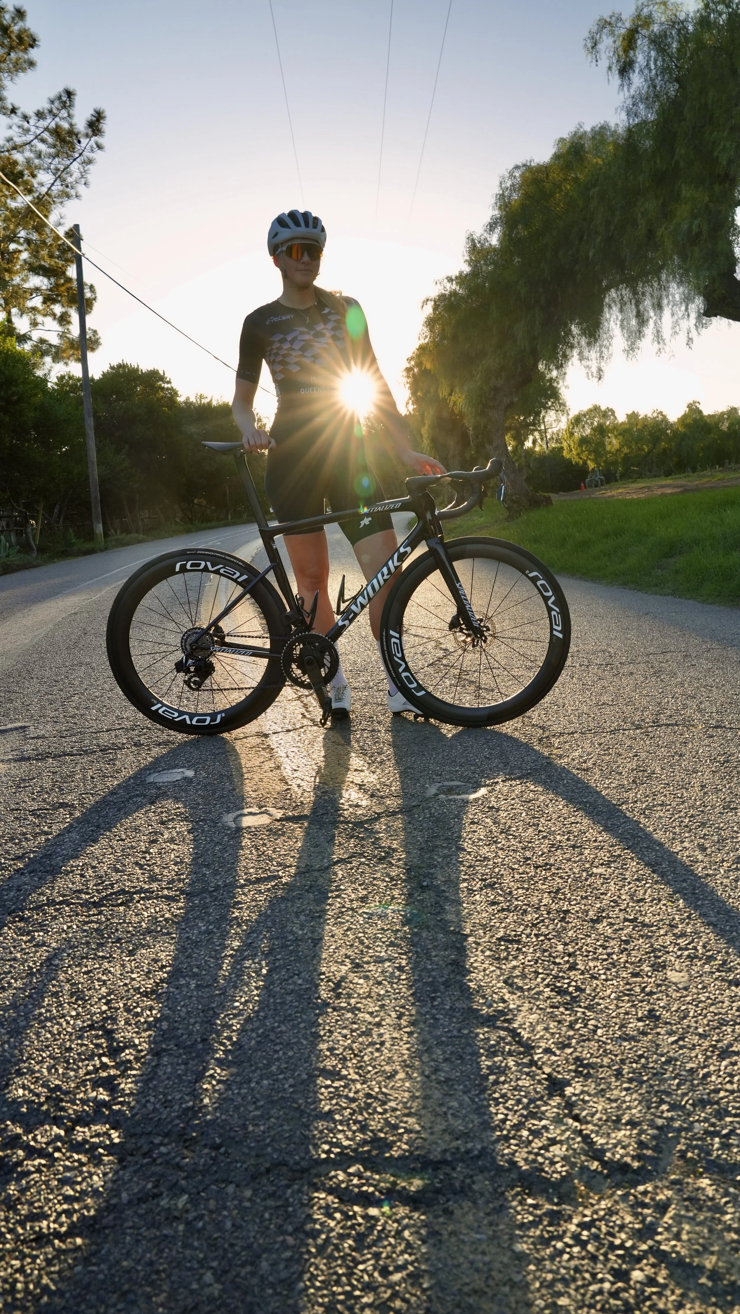 A woman wearing cycling gear and a helmet stands next to a black road bike on a paved road during sunset, with trees on both sides and shadows cast on the ground.
