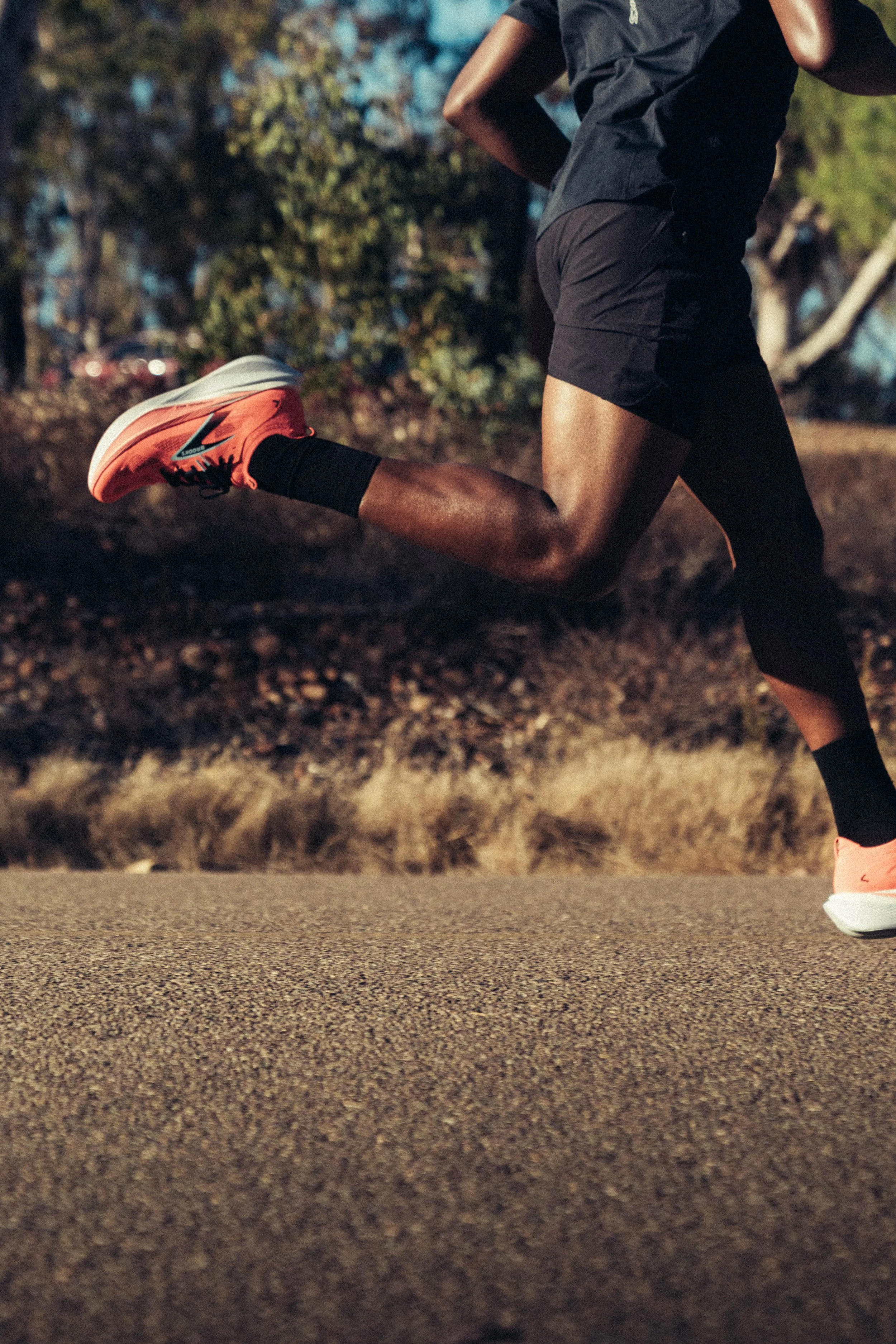 Close-up of a person running outdoors, wearing pink running shoes, black socks, black shorts, and a dark shirt, with trees and blue sky in the background.