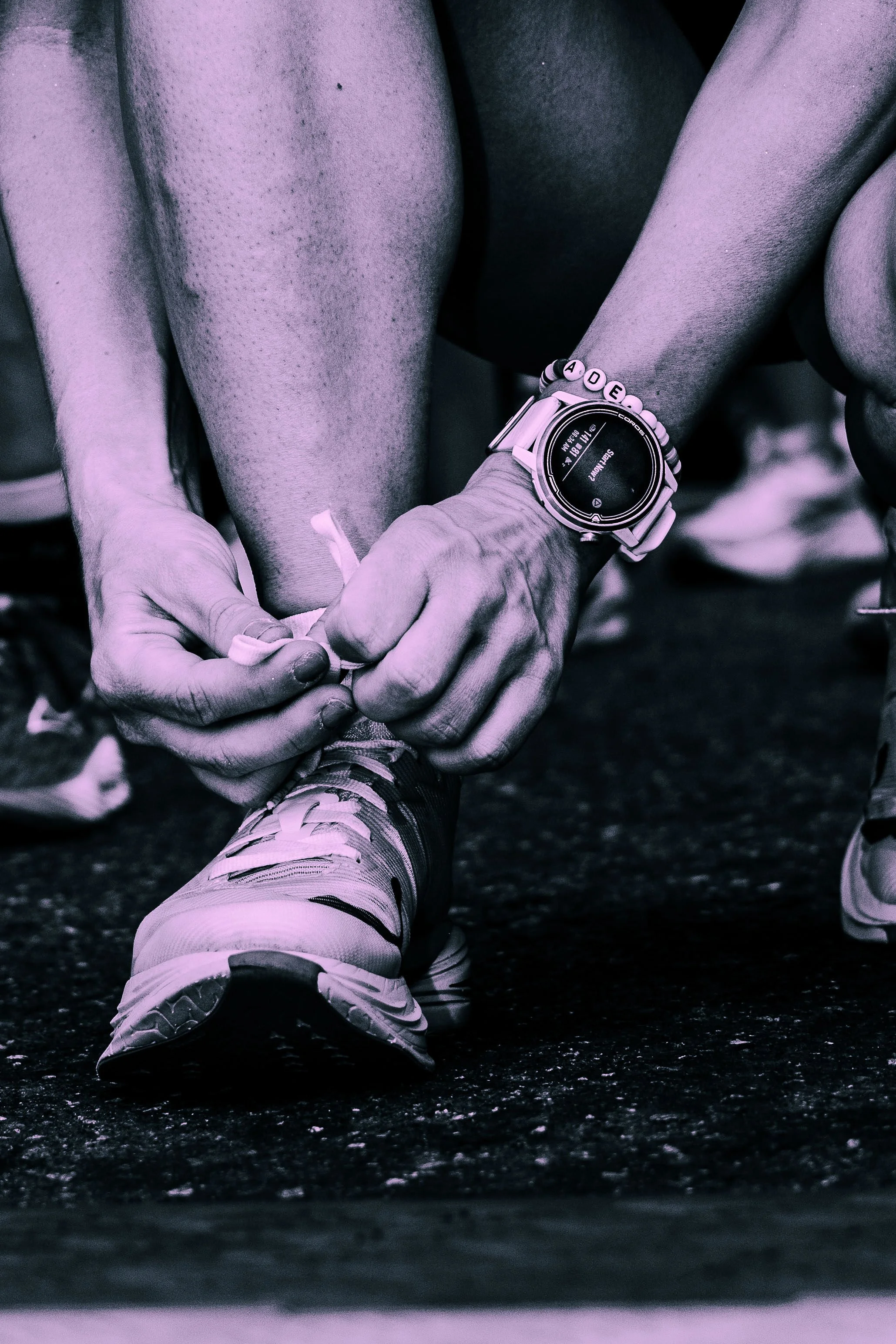 Close-up of a person tying the laces of their running shoes on a track. The person is wearing a smartwatch and a beaded bracelet. The image is in black and white with a hint of purple tint.