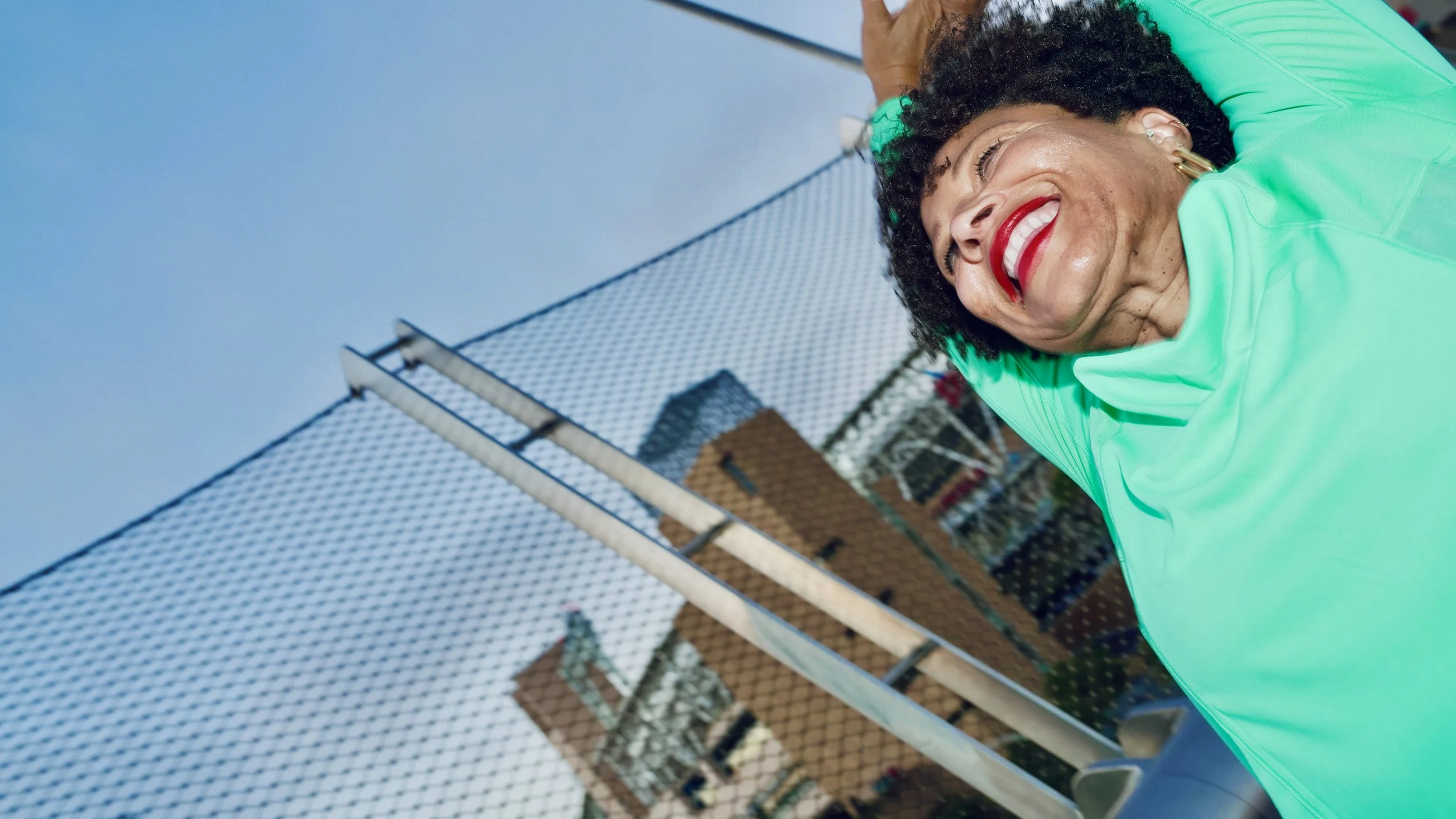 A woman with dark curly hair and red lipstick smiling, wearing a bright teal athletic jacket, standing outdoors near a chain-link fence and a brick building.