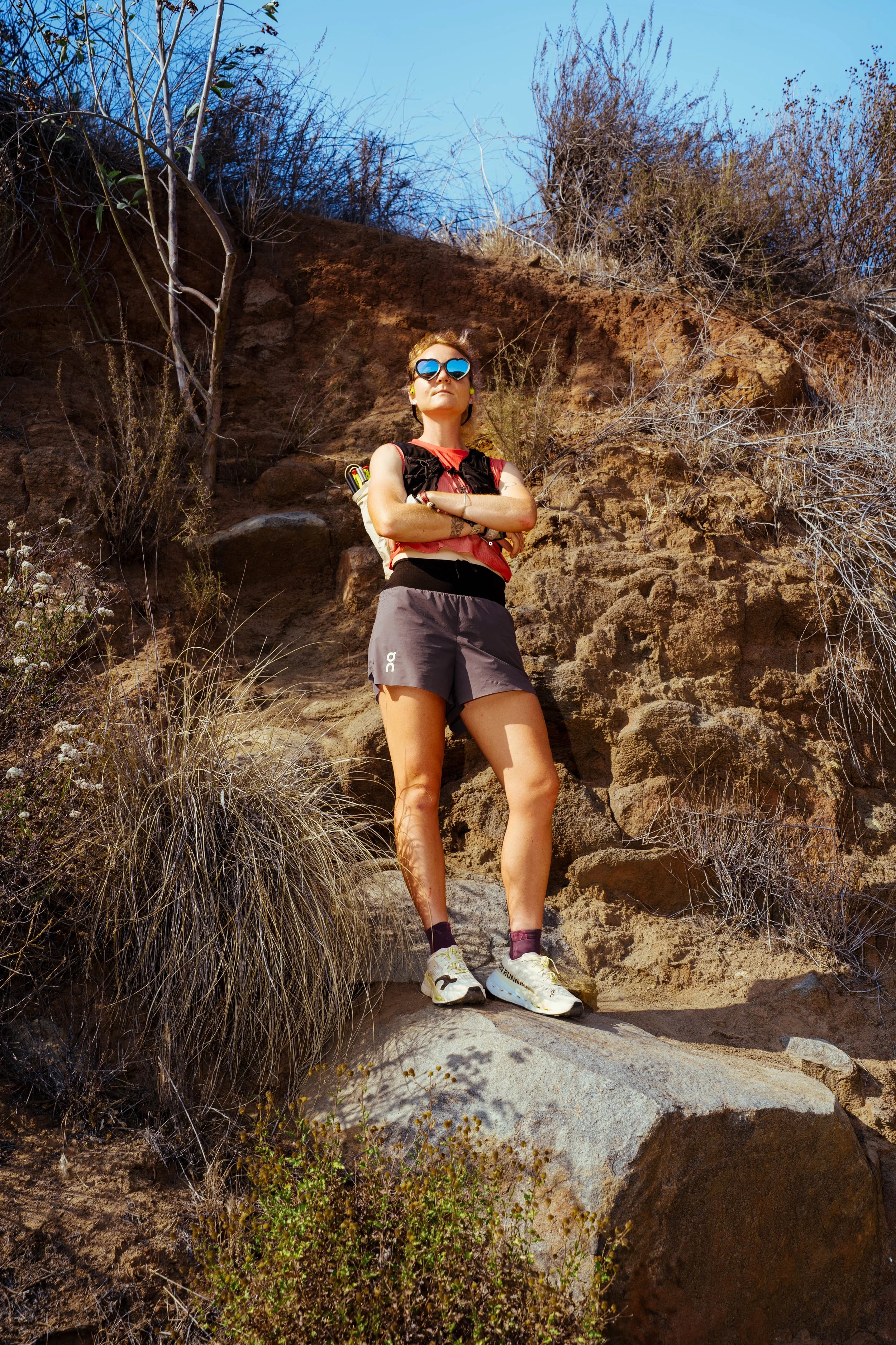 A woman standing on a rocky trail outdoors with arms crossed, wearing sunglasses, a red sleeveless shirt, black shorts, and athletic shoes, surrounded by dry shrubs and rocks under a clear blue sky.