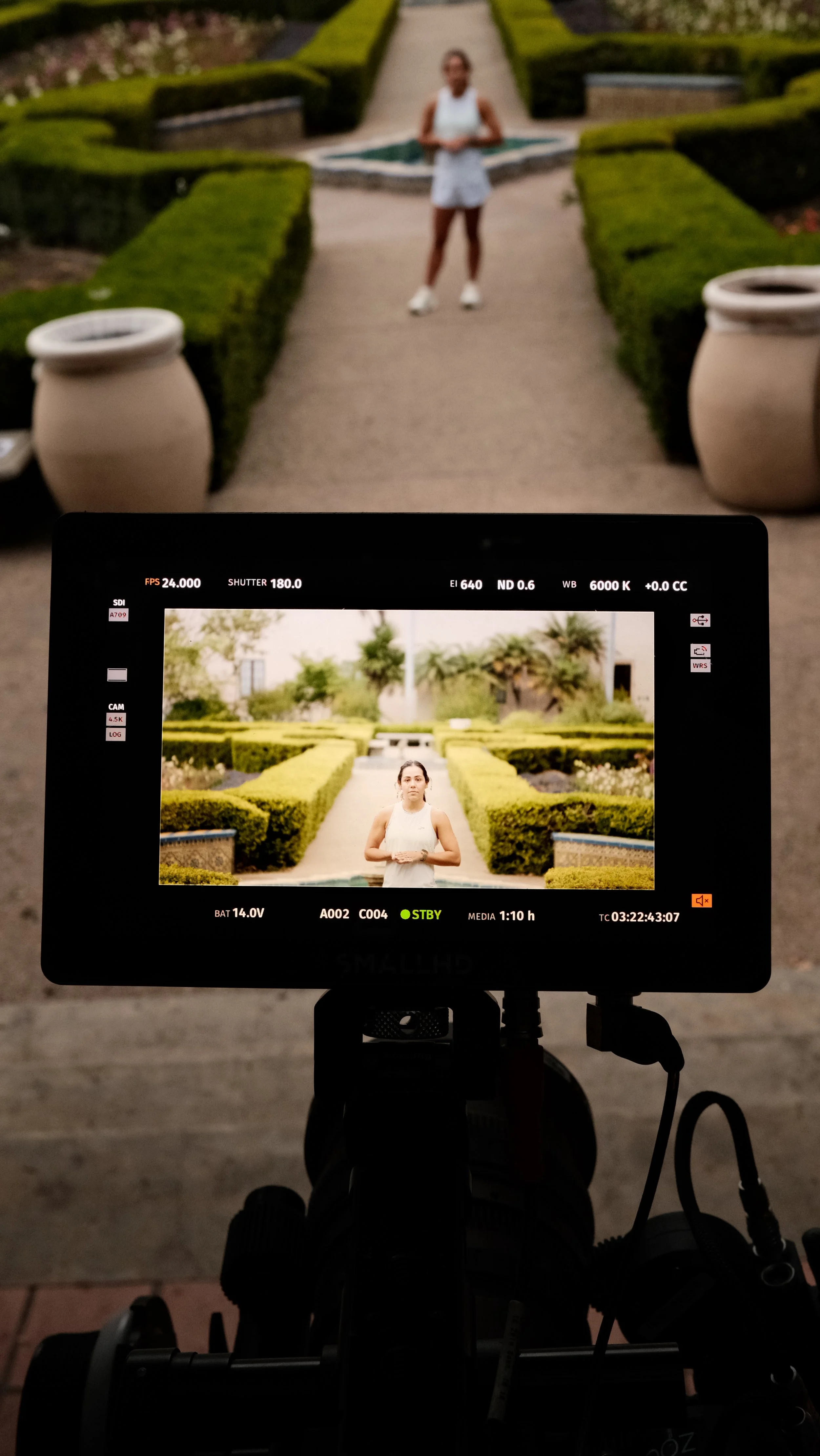A camera monitor displaying a photo of a woman standing in a landscaped garden with hedges and trees, wearing a white sleeveless top and shorts.