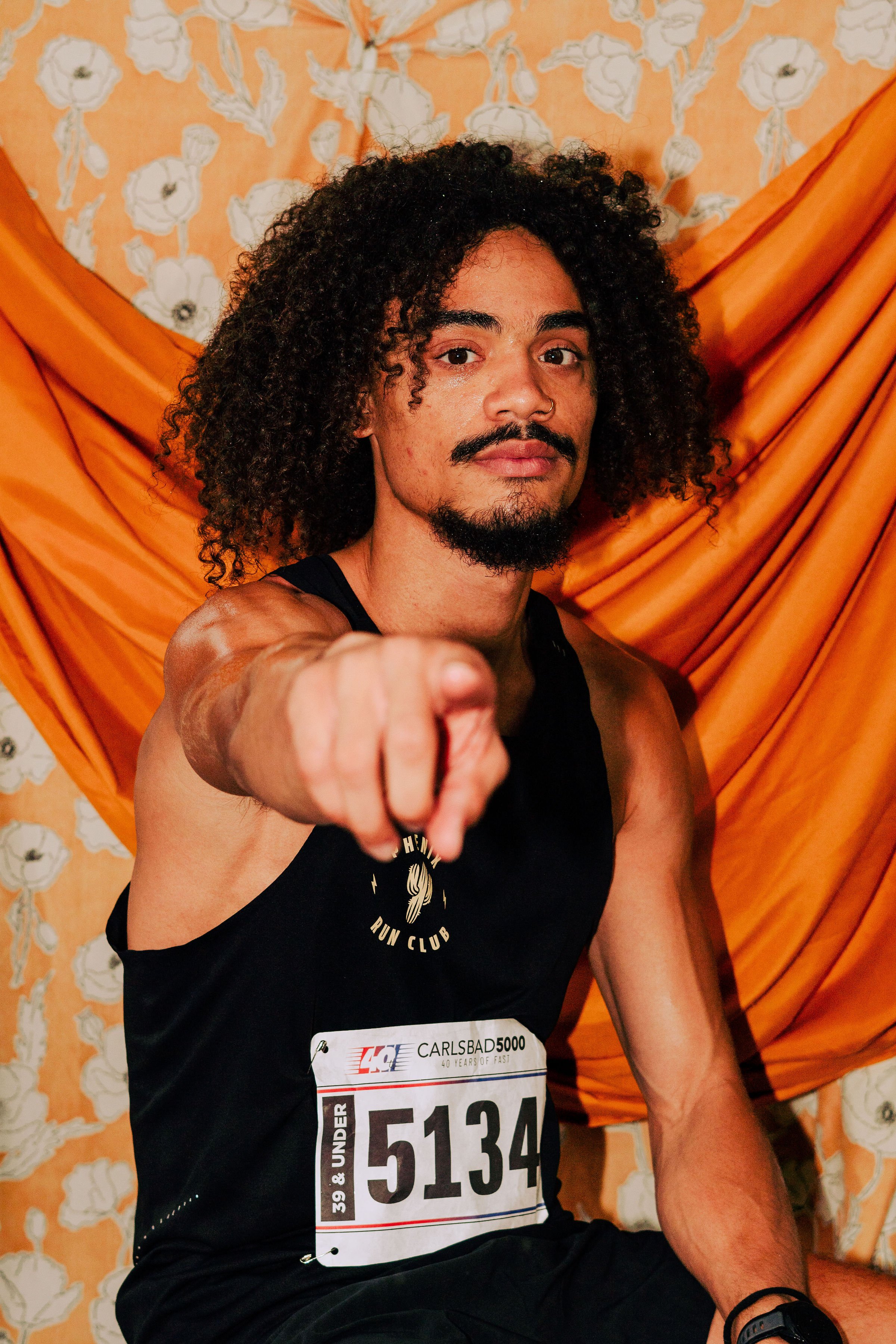 A man with curly hair and a mustache pointing directly at the camera, wearing a black tank top with a race bib, set against an orange patterned fabric background.