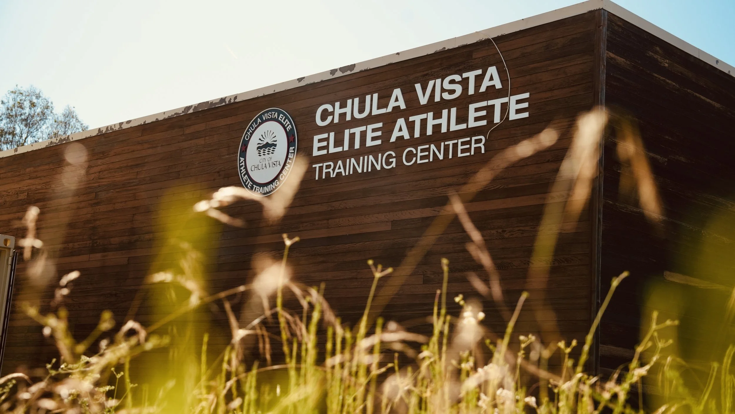 Wooden training center building with signs for Chula Vista Elite Athlete Training Center, viewed through grass with trees and a blue sky in the background.