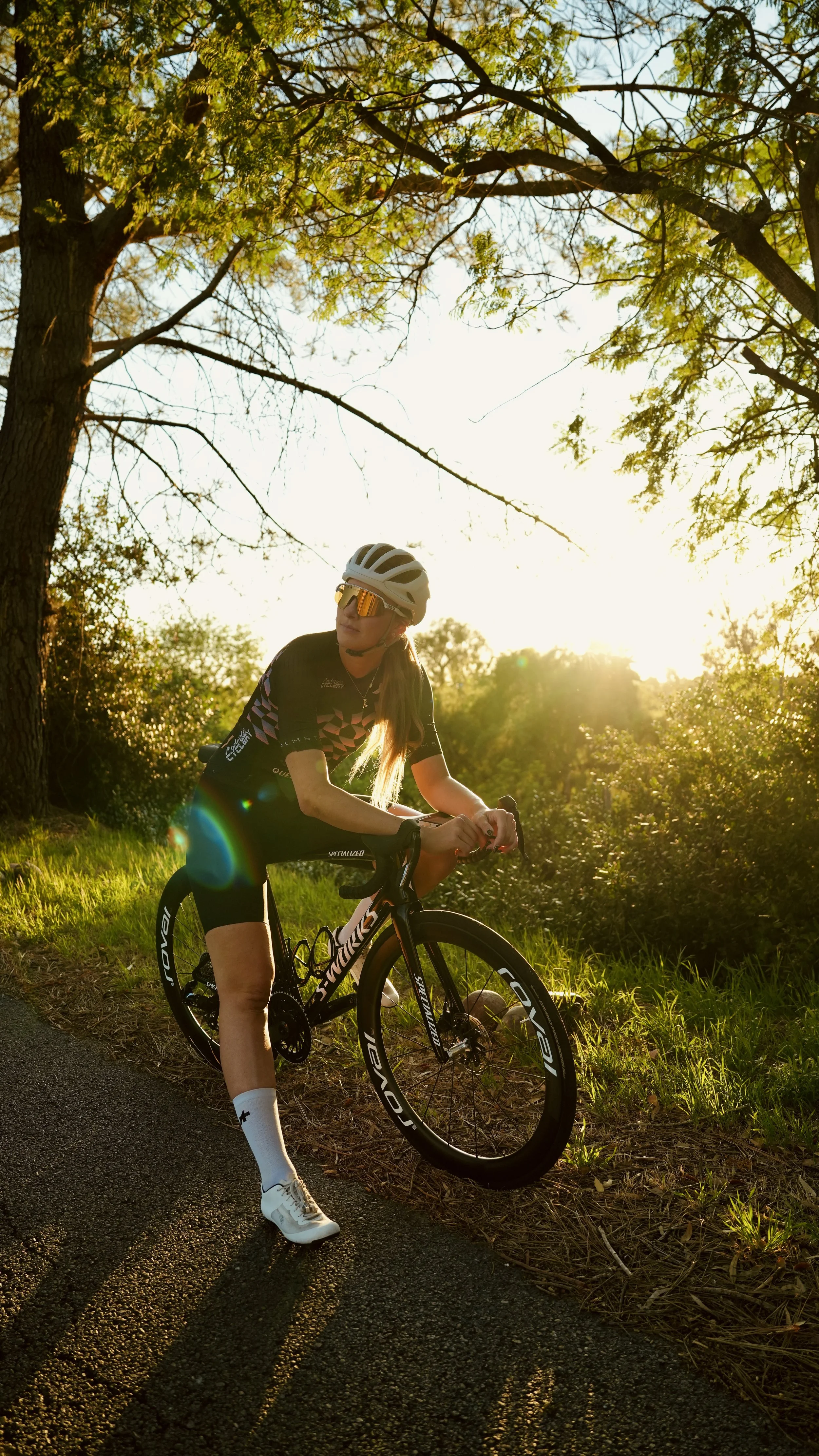 A woman wearing a helmet, sunglasses, cycling jersey, cycling shorts, and socks is standing with her black gravel bike on a paved path next to grass and bushes, under trees, with the sun setting in the background.