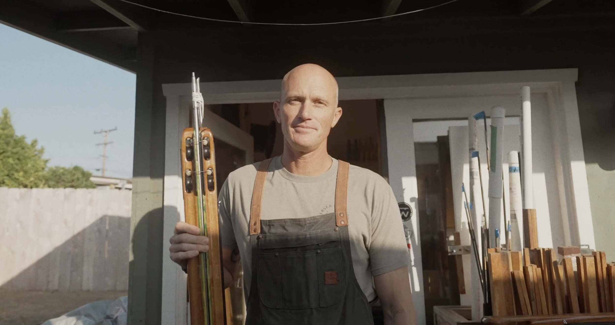 A man standing outdoors in front of a building, holding a pair of skis, with other ski poles and equipment nearby.