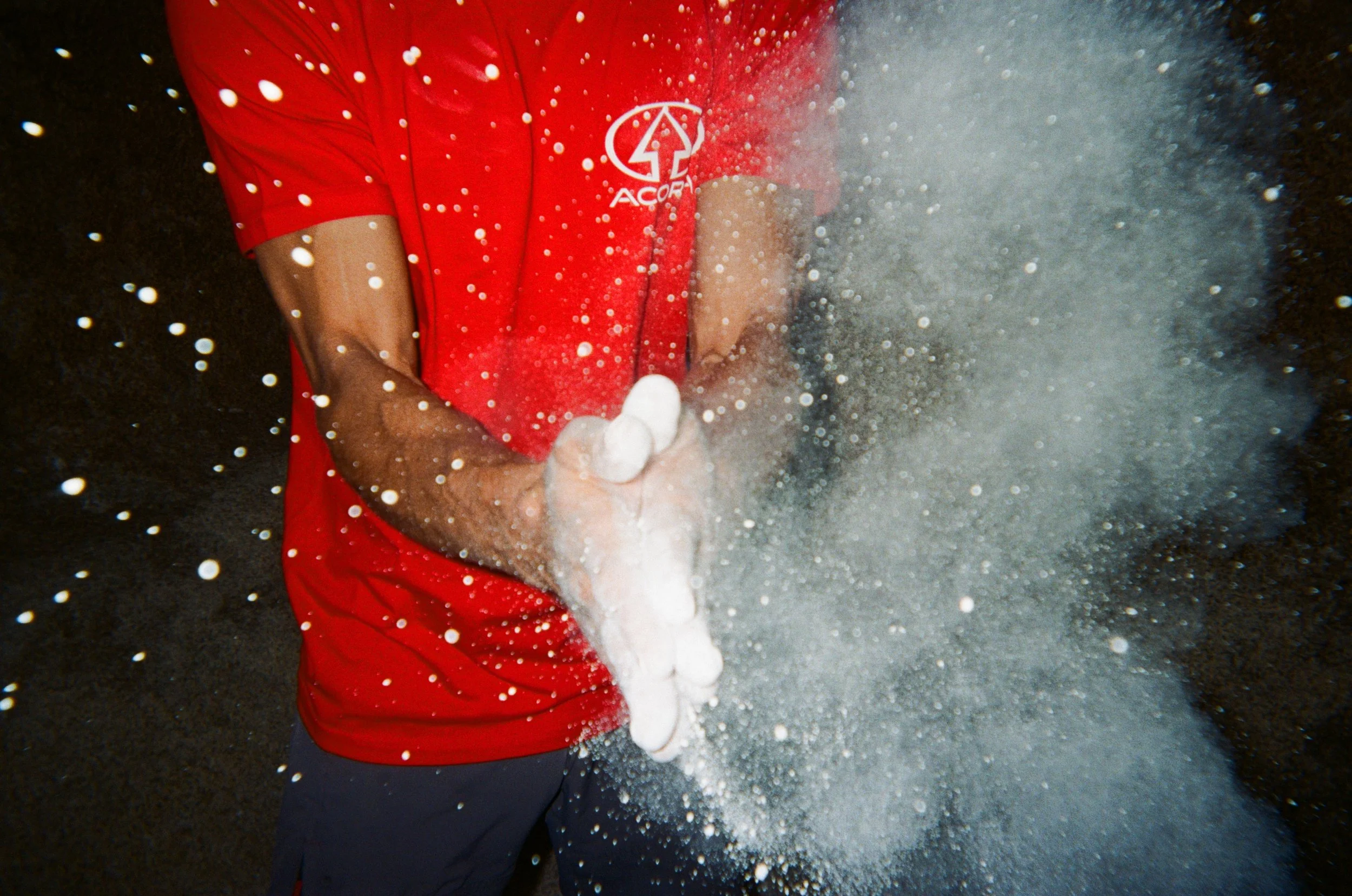 A person wearing a red shirt with a white logo on it, standing in front of a dark background, creating a cloud of white foam or spray with their hands.