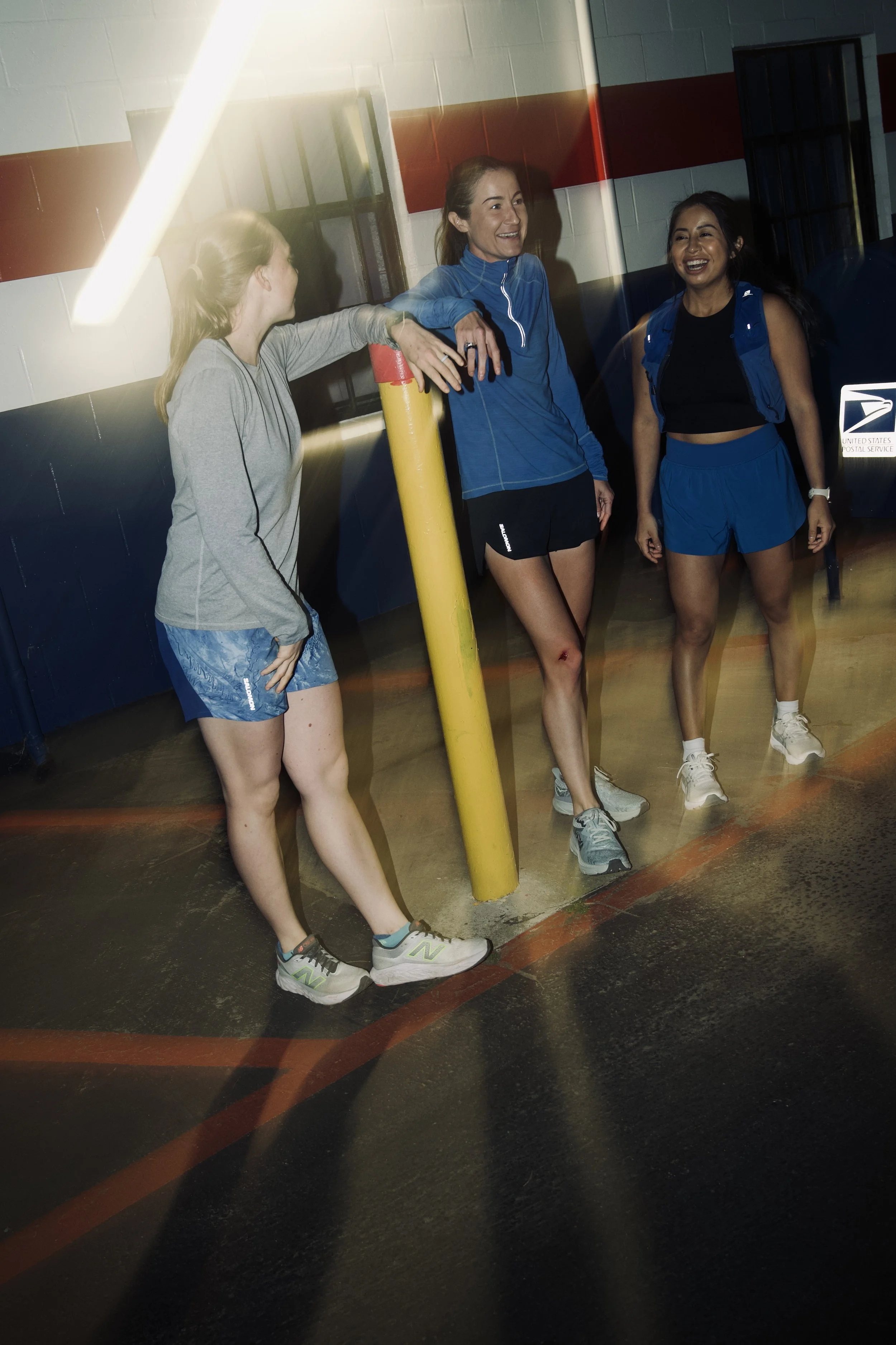 Three women in athletic clothing standing near a yellow post in a gym, smiling and engaging in conversation.