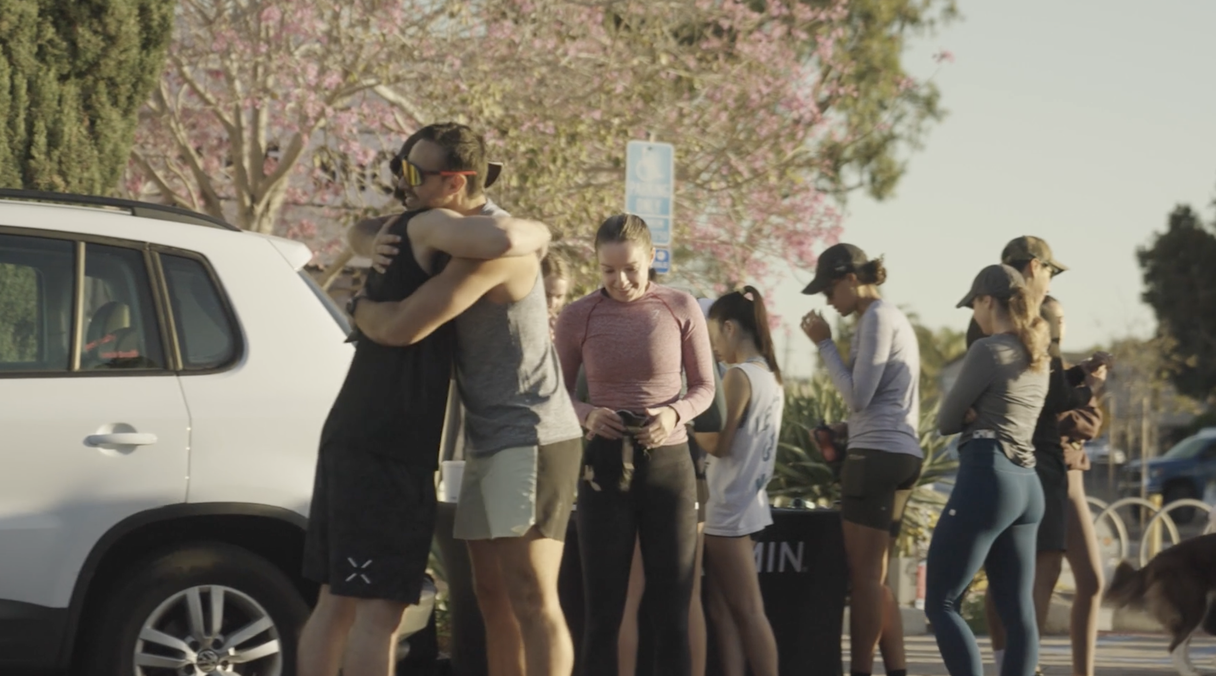 People standing in line outdoors on a sunny day, with pink flowering trees in the background.