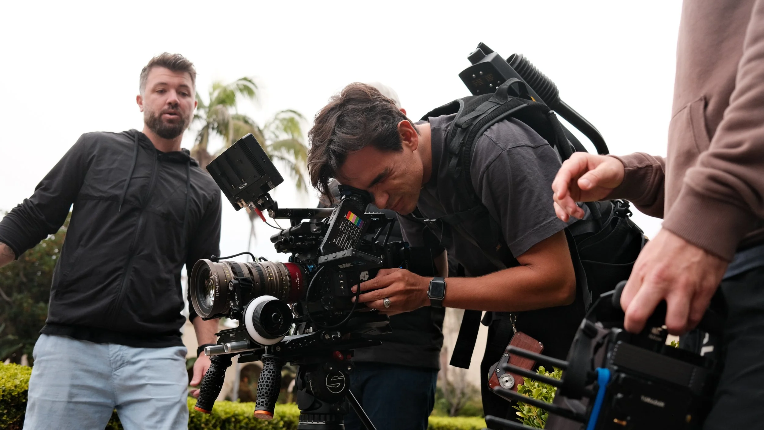 Group of people working with professional video camera outdoors, with palm trees in the background.