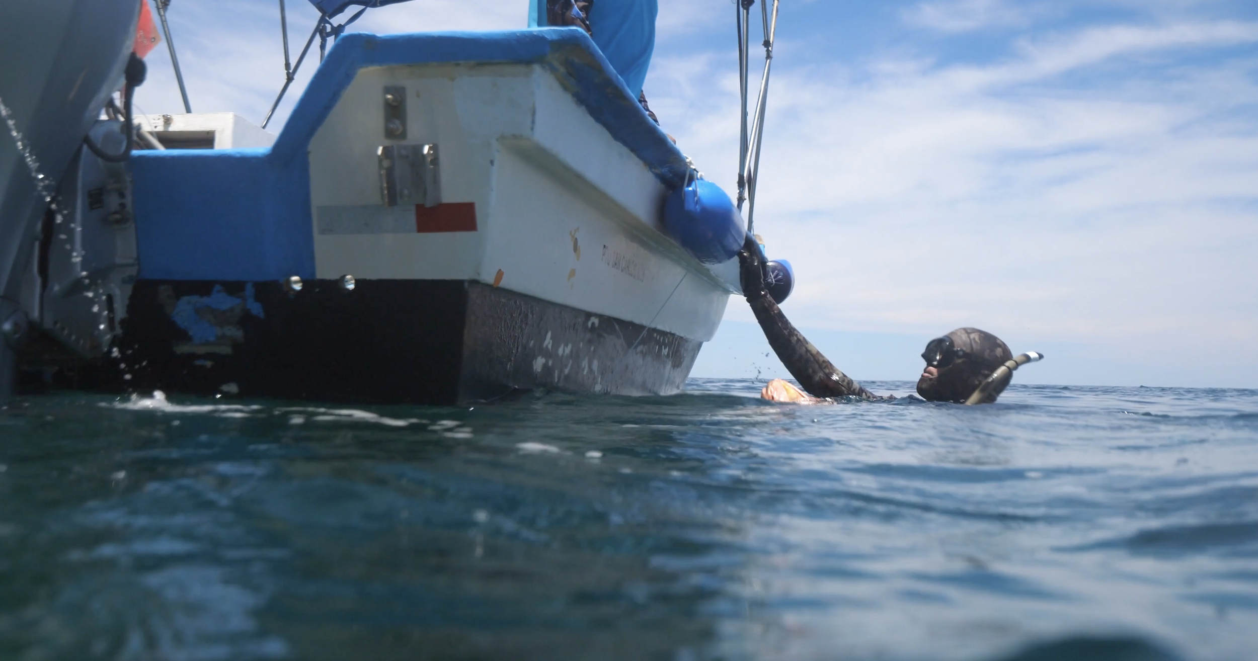 A diver swimming in the water next to a boat, holding onto the boat's side with one hand, wearing a diving mask and wetsuit, with the sky and ocean in the background.