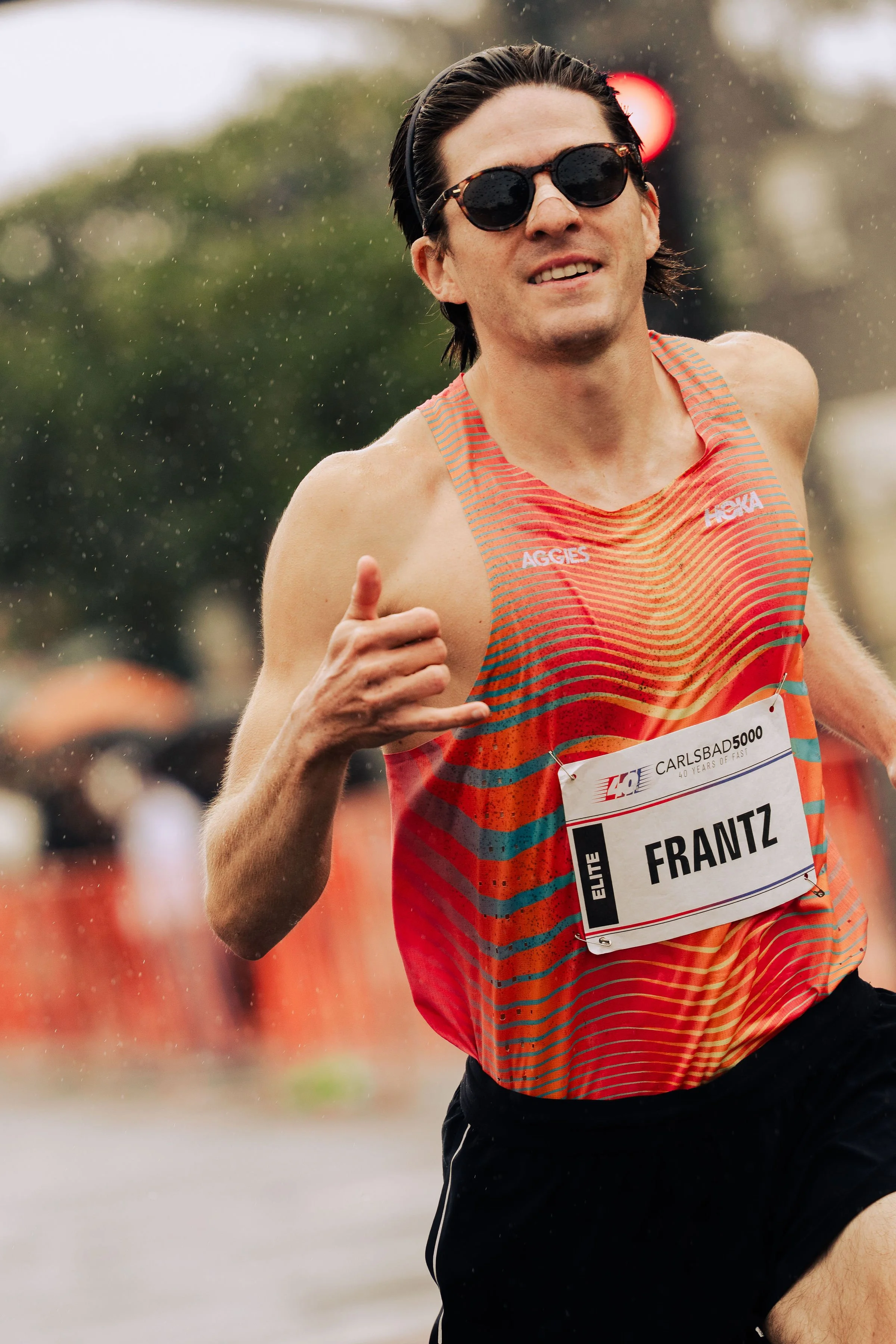 A male runner with sunglasses, wet hair, wearing an orange striped running outfit, smiling and giving a thumbs-up during a race.