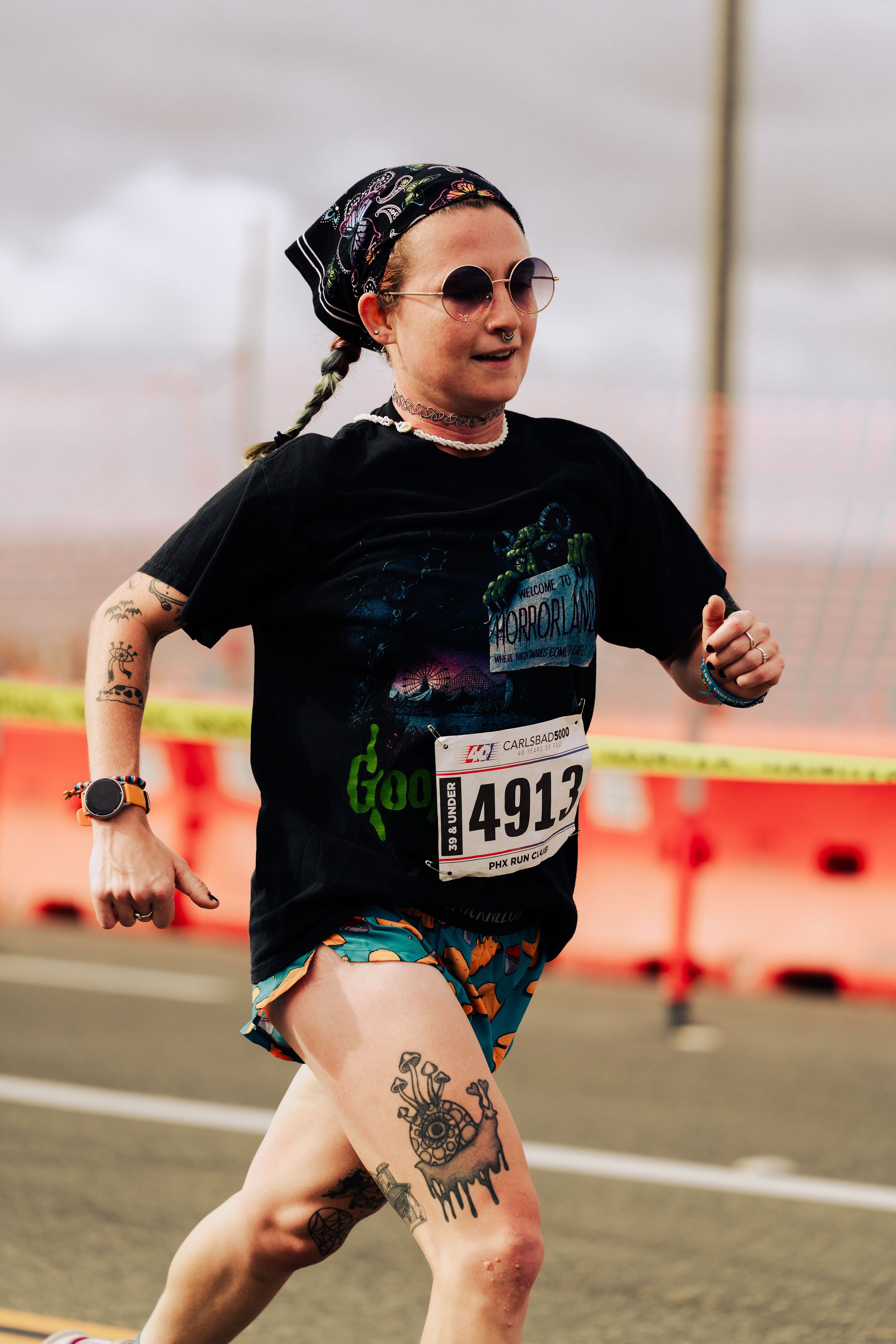 A woman running on a track during a race, wearing sunglasses, a black t-shirt with a graphic, colorful shorts, and sporting tattoos on her arm and leg. She has a headscarf, jewelry, and a race bib numbered 4913.