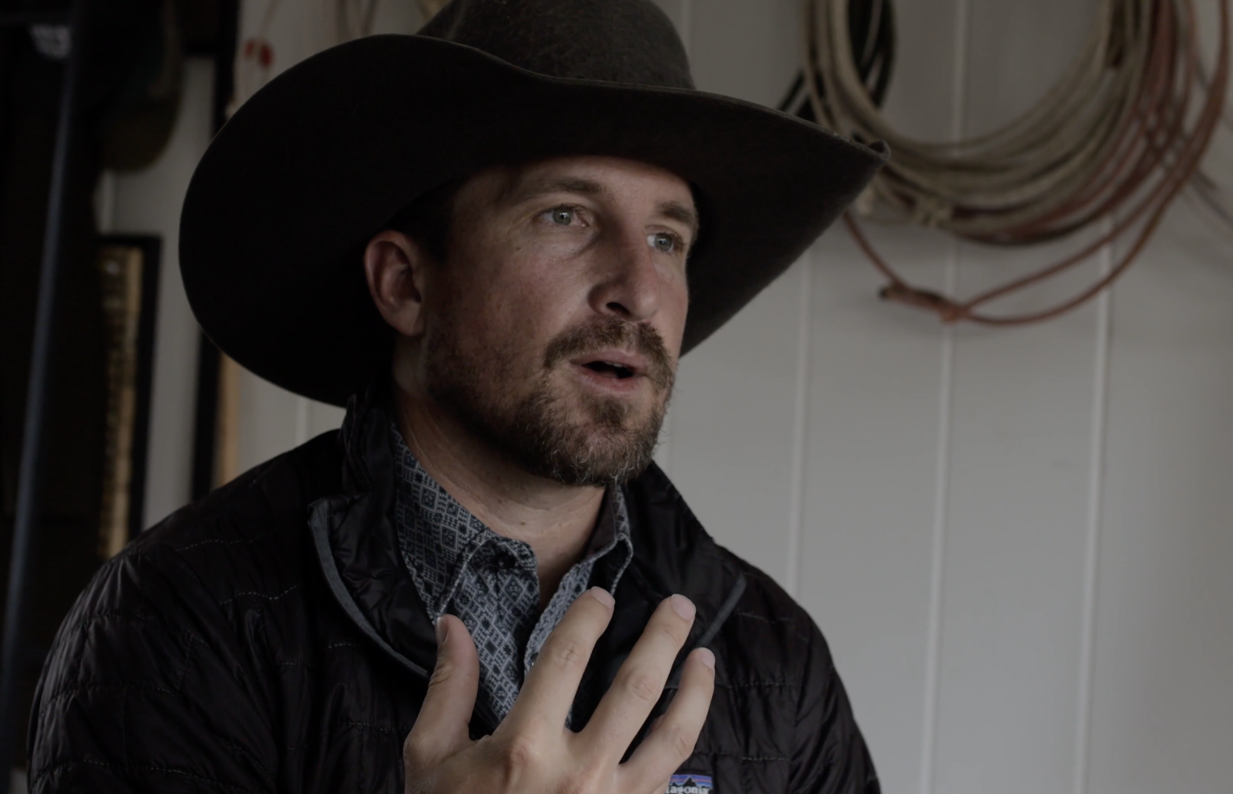 A man wearing a cowboy hat and a black jacket, with his hand over his chest, appears to be speaking or singing, indoors with a coiled hose on the wall behind him.