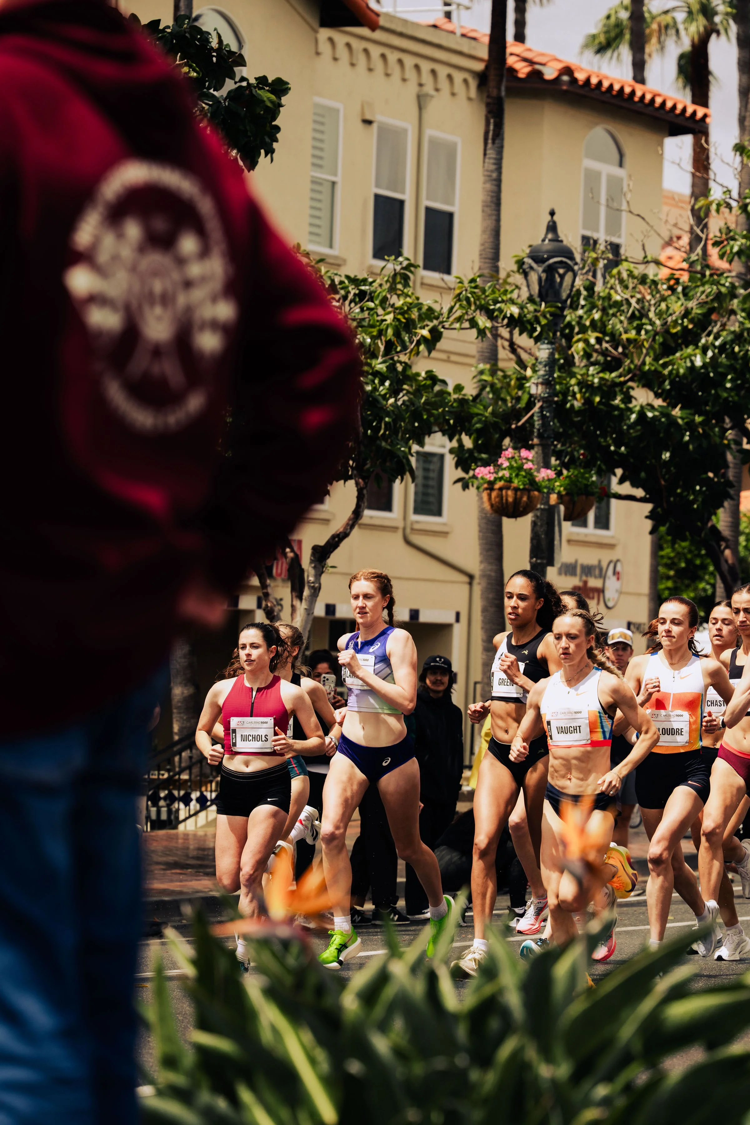 A group of women participating in a marathon on a city street, with buildings, trees, and hanging flower baskets in the background.