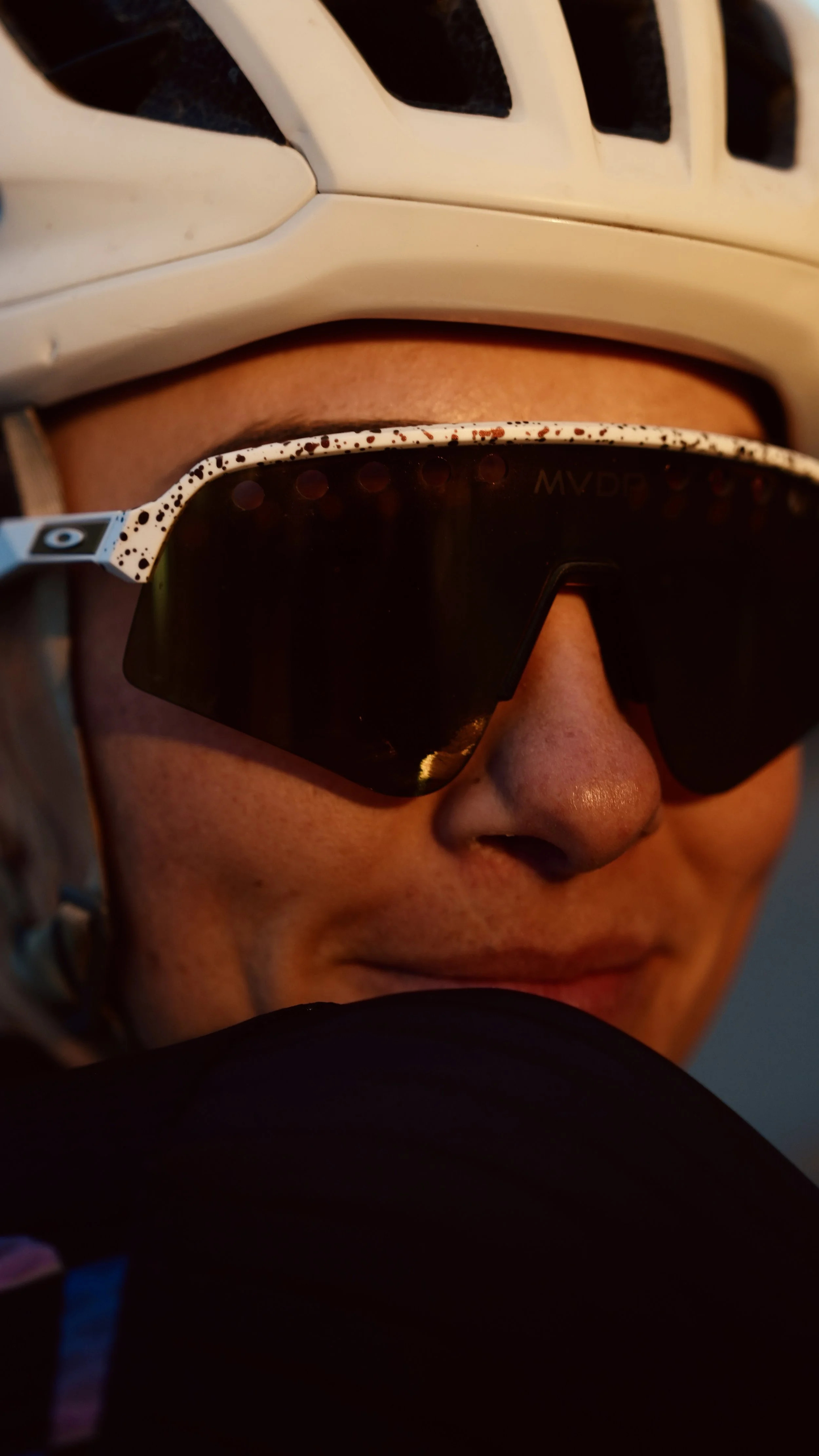 Close-up of a person wearing white sports sunglasses with black speckles and a white helmet. The person has a tan complexion and is partially obscured by a black object in the foreground.