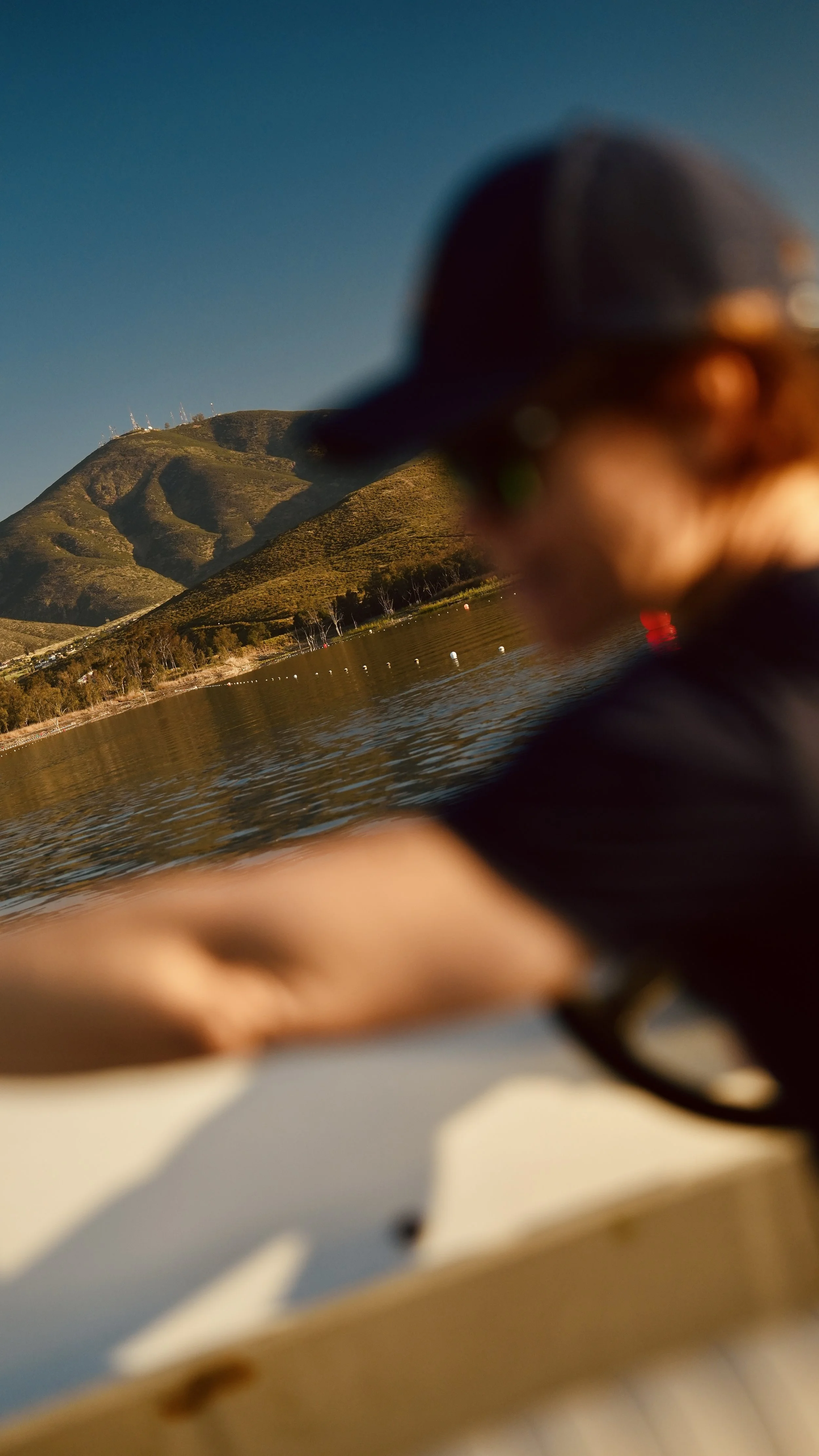 Blurred view of a mountain with some wind turbines on top, reflected in a body of water, with part of a person's hand and a watch in the foreground.