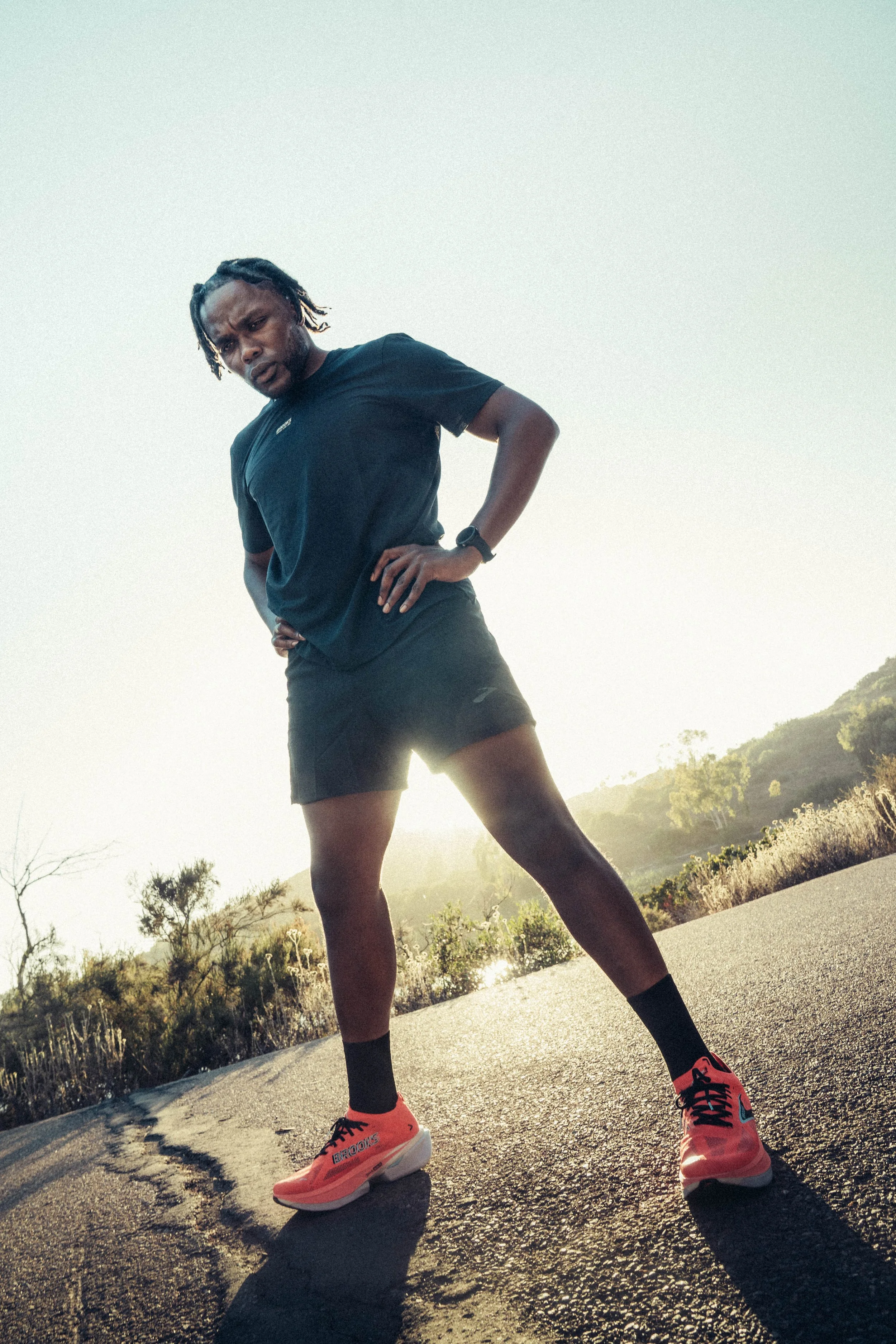 Male athlete in black athletic wear, orange running shoes, standing on a road with hands on hips, during sunset with hills and trees in the background.