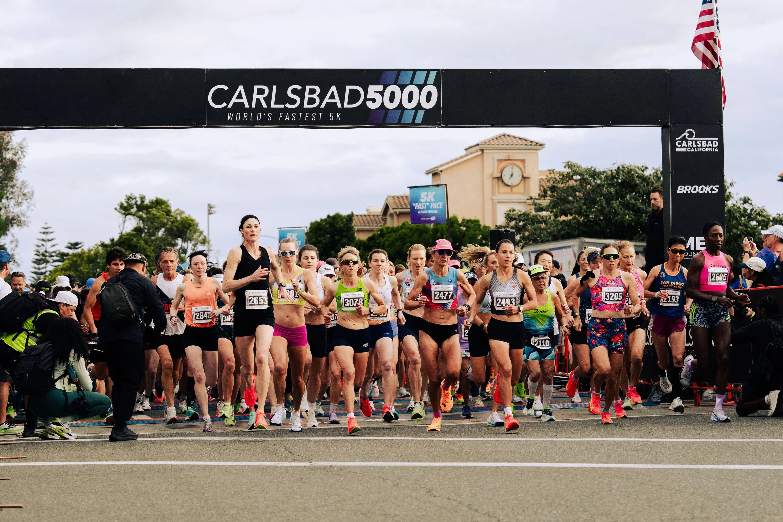 Group of people participating in a marathon or race at the starting line, with a large black banner overhead reading 'Carlsbad 5000, World's Fastest 5K.' Some runners are wearing athletic gear and numbered bibs, and there are spectators and officials