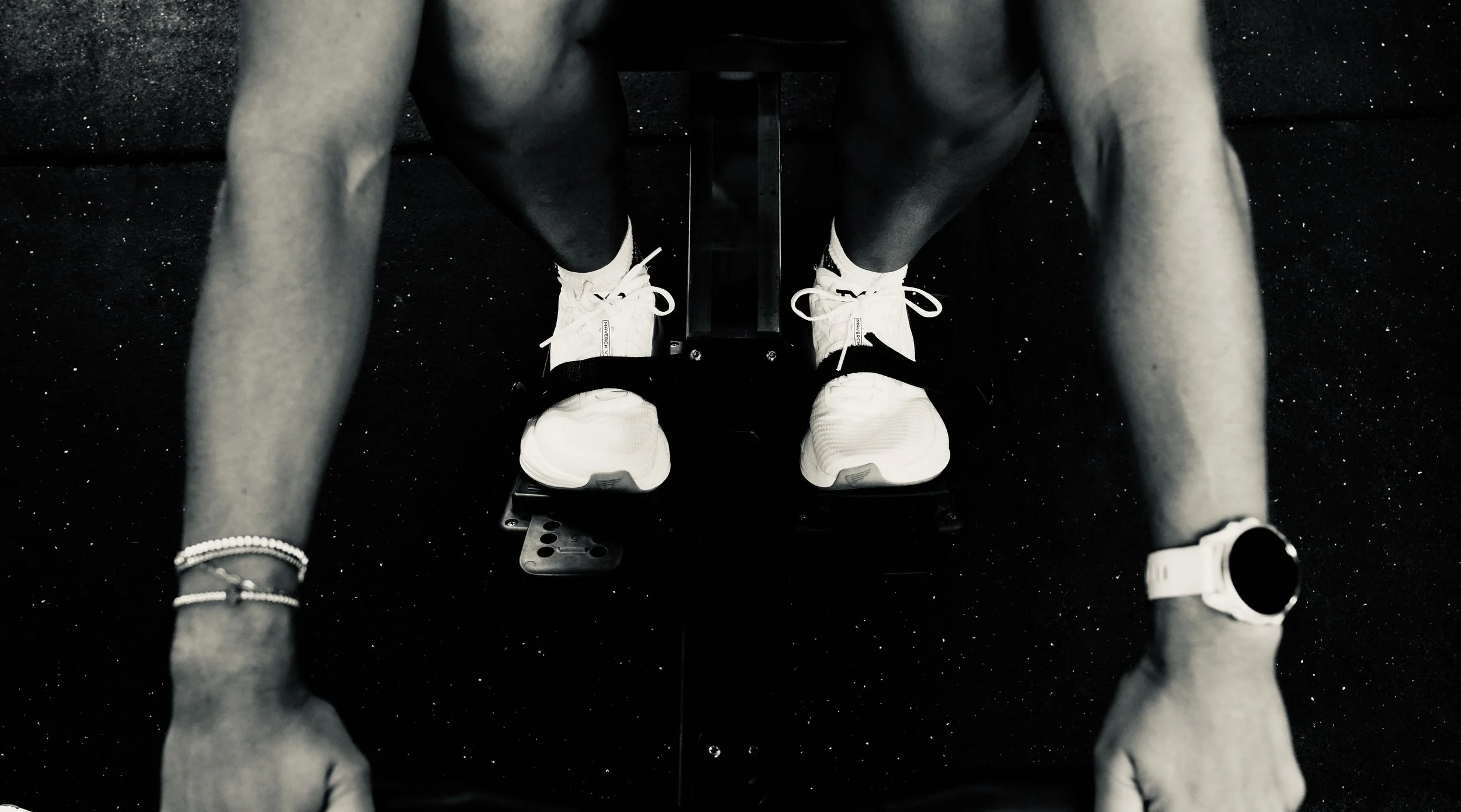 Close-up of a person wearing white sneakers and a white sports watch, doing push-ups on a black speckled gym floor.