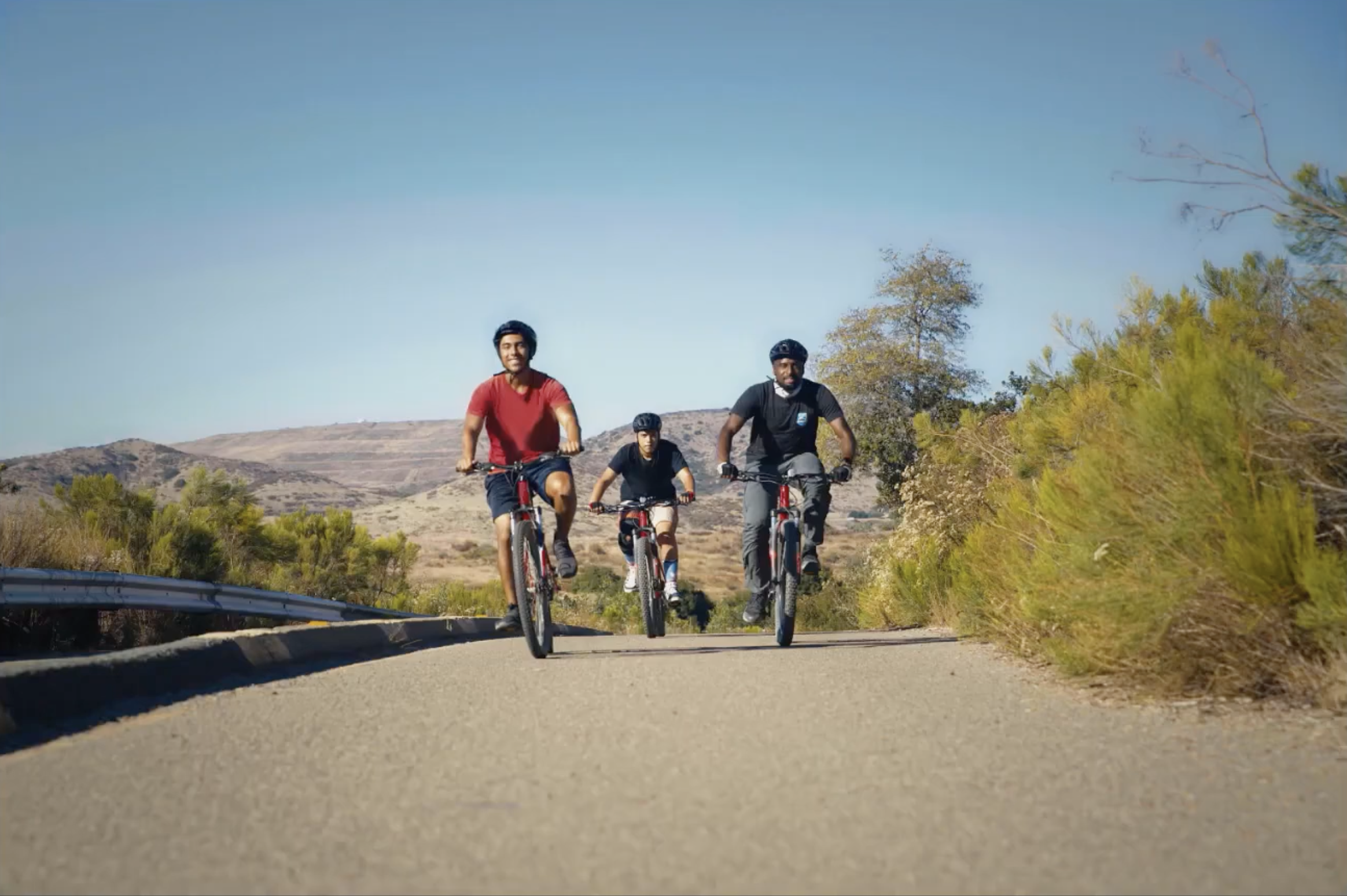 Four people riding mountain bikes on a sunny outdoor trail with hills and trees in the background.