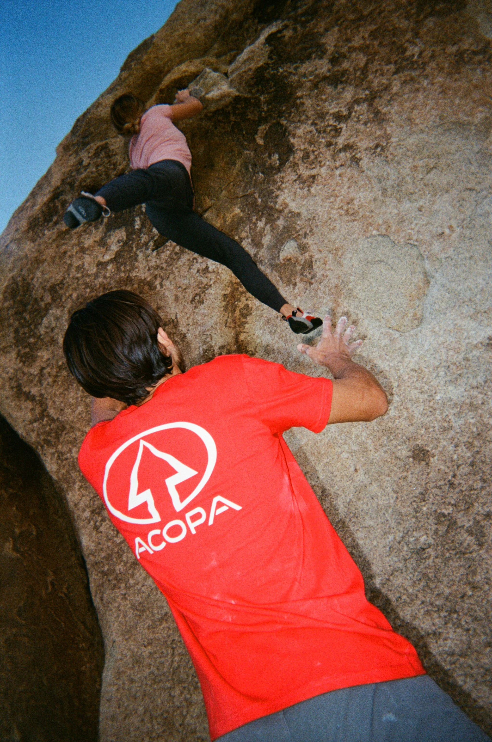 A person climbing a large rock face outdoors, wearing a pink shirt and black pants, with another person spotting at the base.