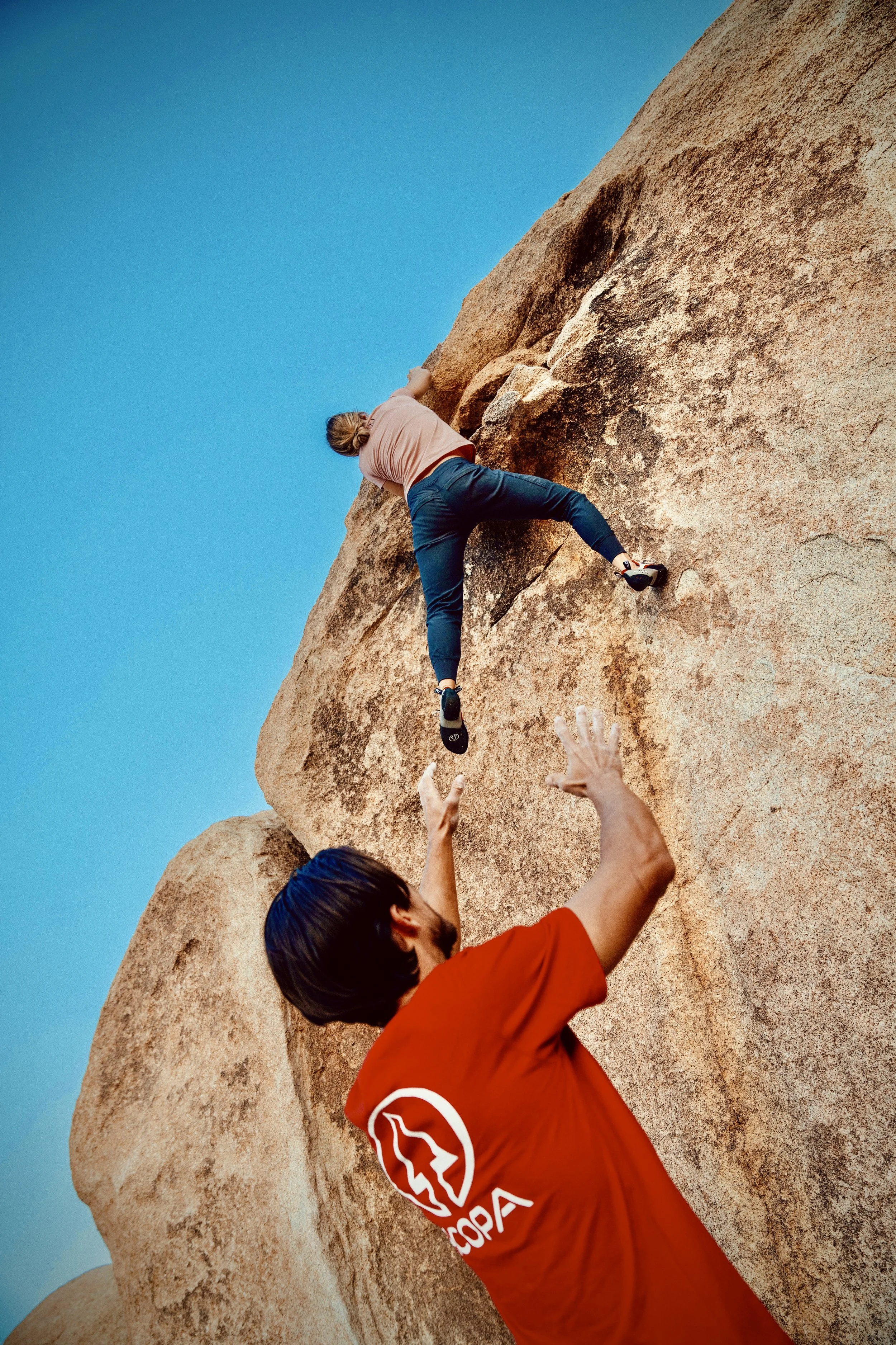 A person in a pink shirt and blue pants is rock climbing on a large granite boulder with helper hands beneath them, showing they are being assisted or spotted during the climb, with a clear blue sky in the background.