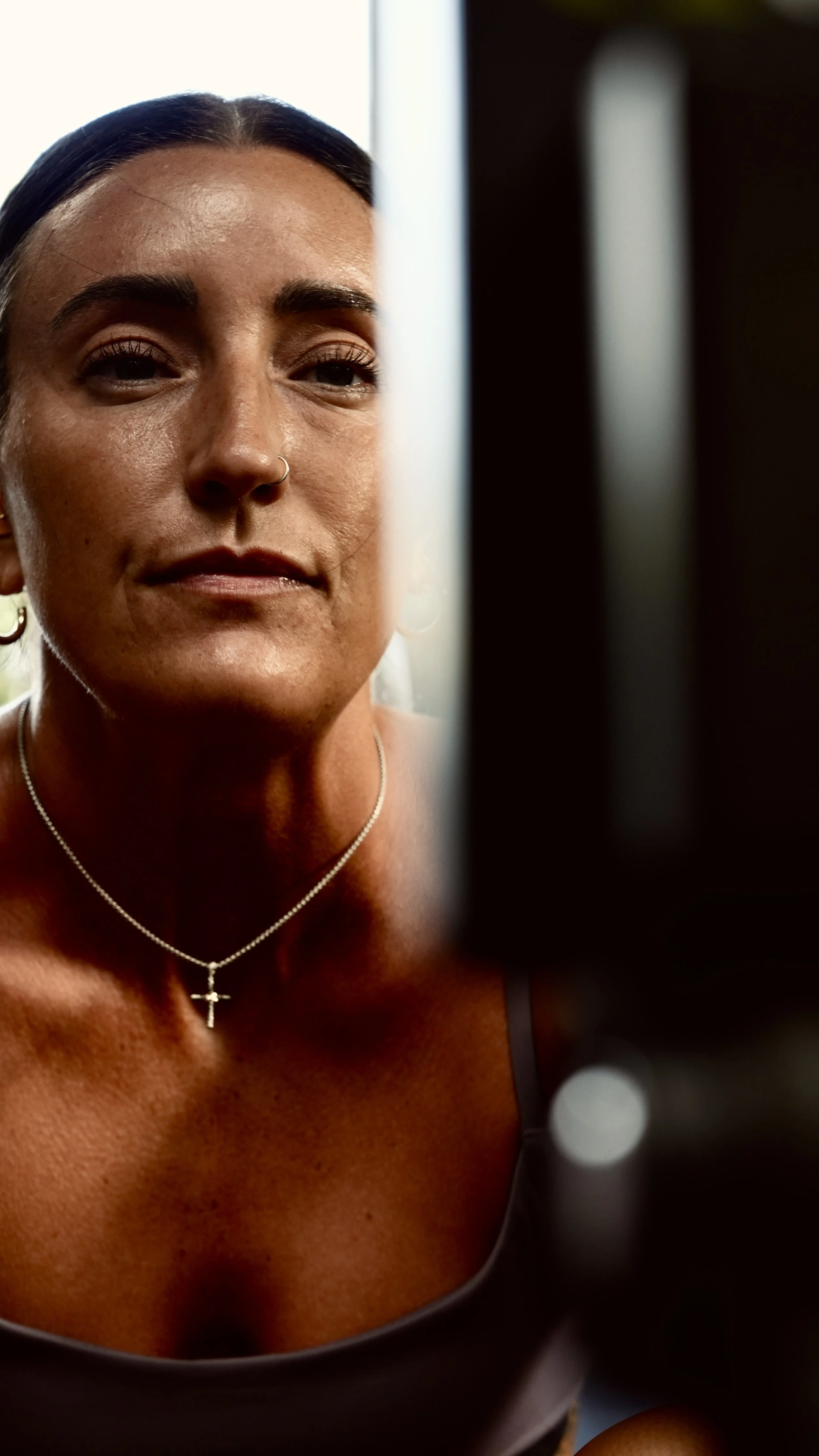 Close-up of a woman with dark hair, tan skin, wearing a silver cross necklace, black tank top, with a serious or neutral expression, standing near a mirror or window with light behind her.