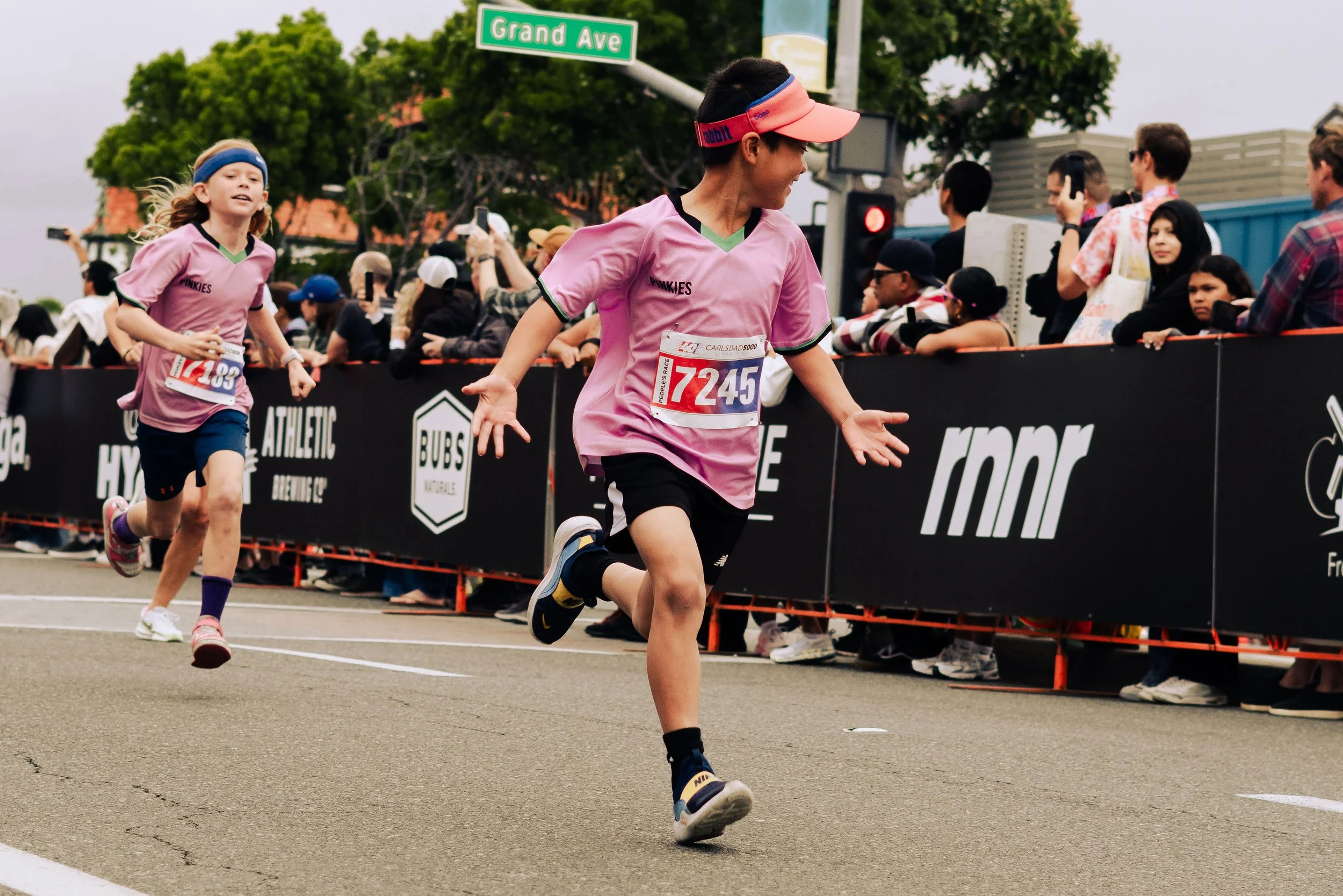 Two children running in a race at an outdoor event, wearing pink t-shirts and racing bibs, with spectators watching from behind a barrier.