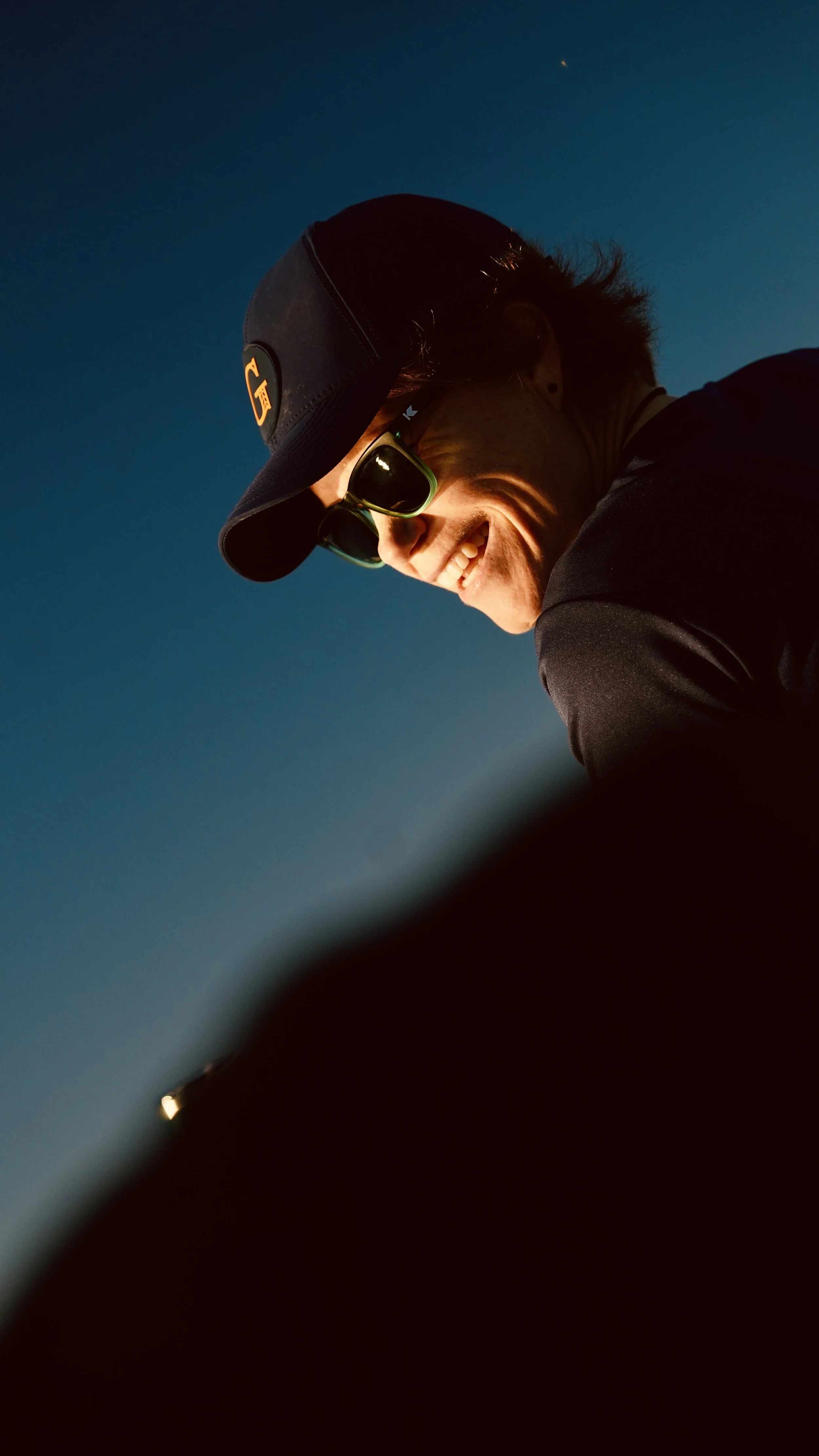 A person wearing a black cap, sunglasses, and dark clothing, smiling against a clear blue sky with a faint star or bright object visible in the distance.