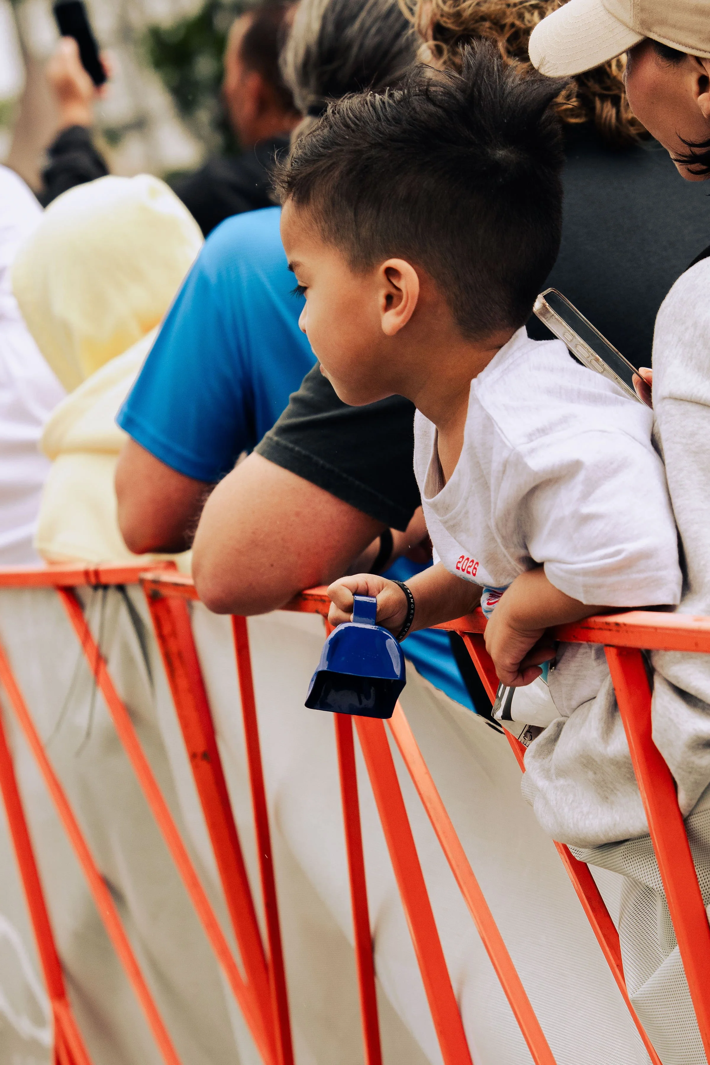 People standing behind an orange metal barrier, with some holding a phone and others holding a water bottle. The focus is on a young person with short black hair, wearing a white shirt, leaning on the barrier.