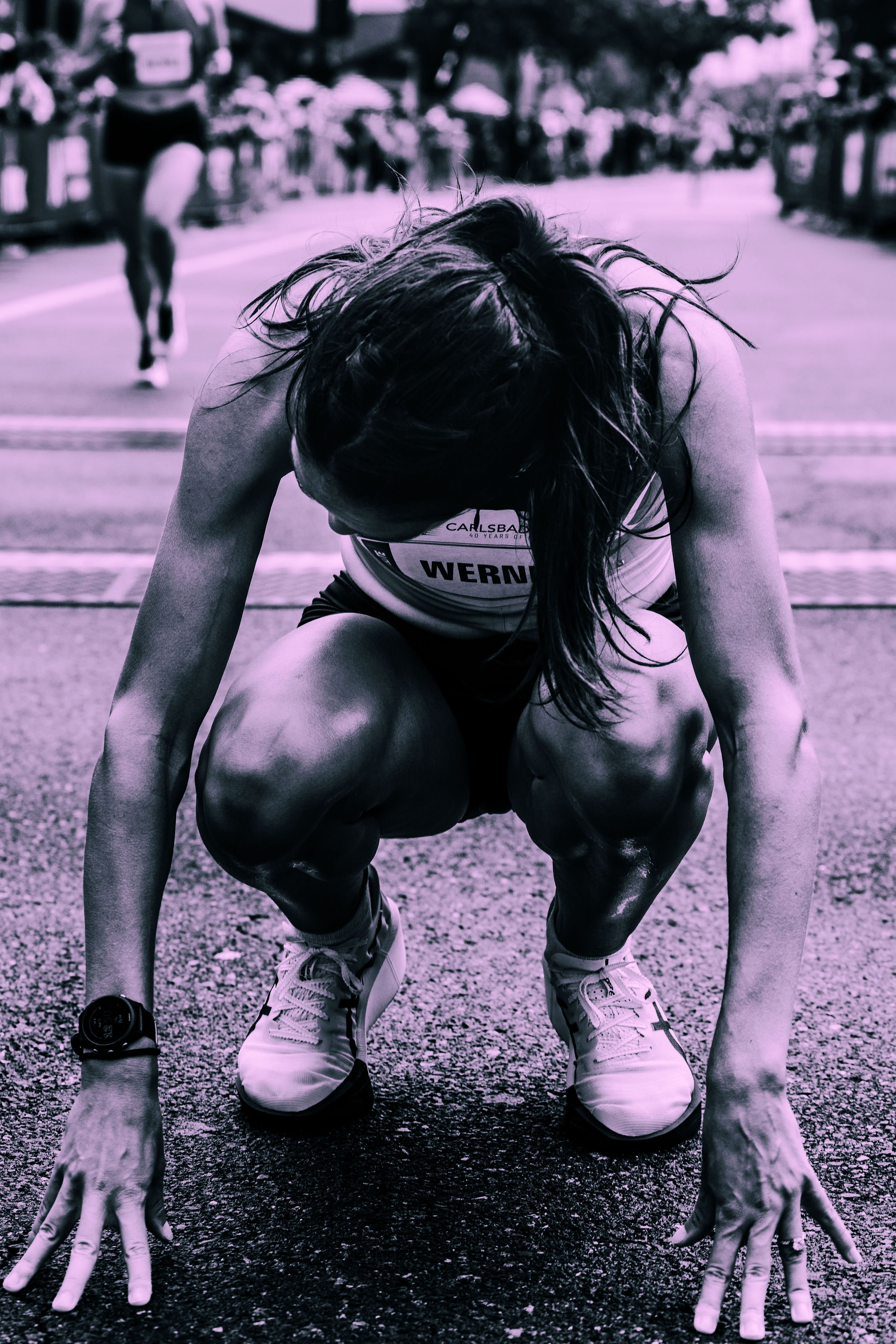 A female athlete in a crouched starting position wearing running gear, including a bib with the name 'Werner' and a race watch on her wrist, on a track in a race setting with spectators in the background.
