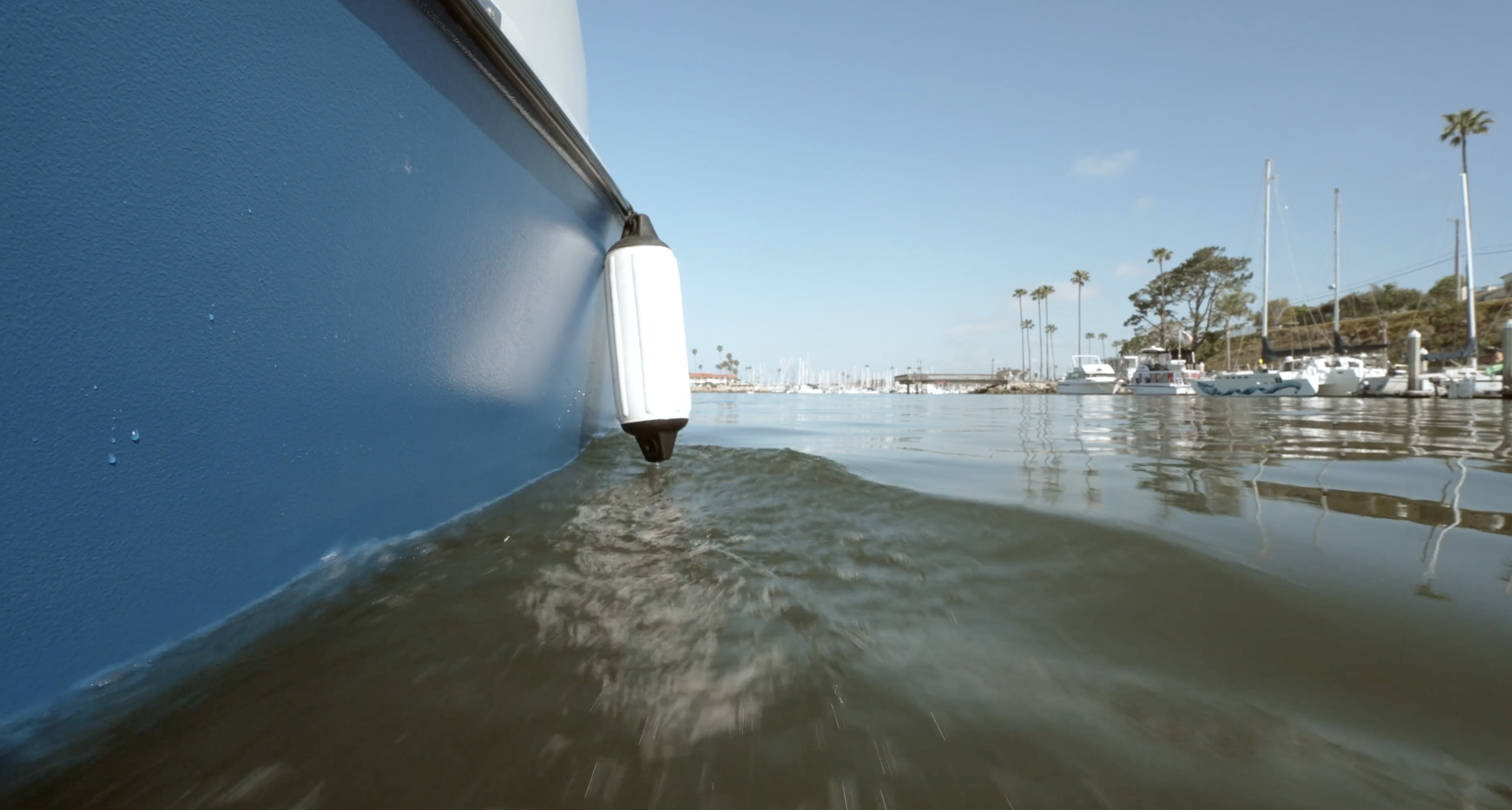 Close-up of a blue boat's hull with a fender hanging over the side, reflected in calm marina waters, with white sailboats, palm trees, and a clear blue sky in the background.
