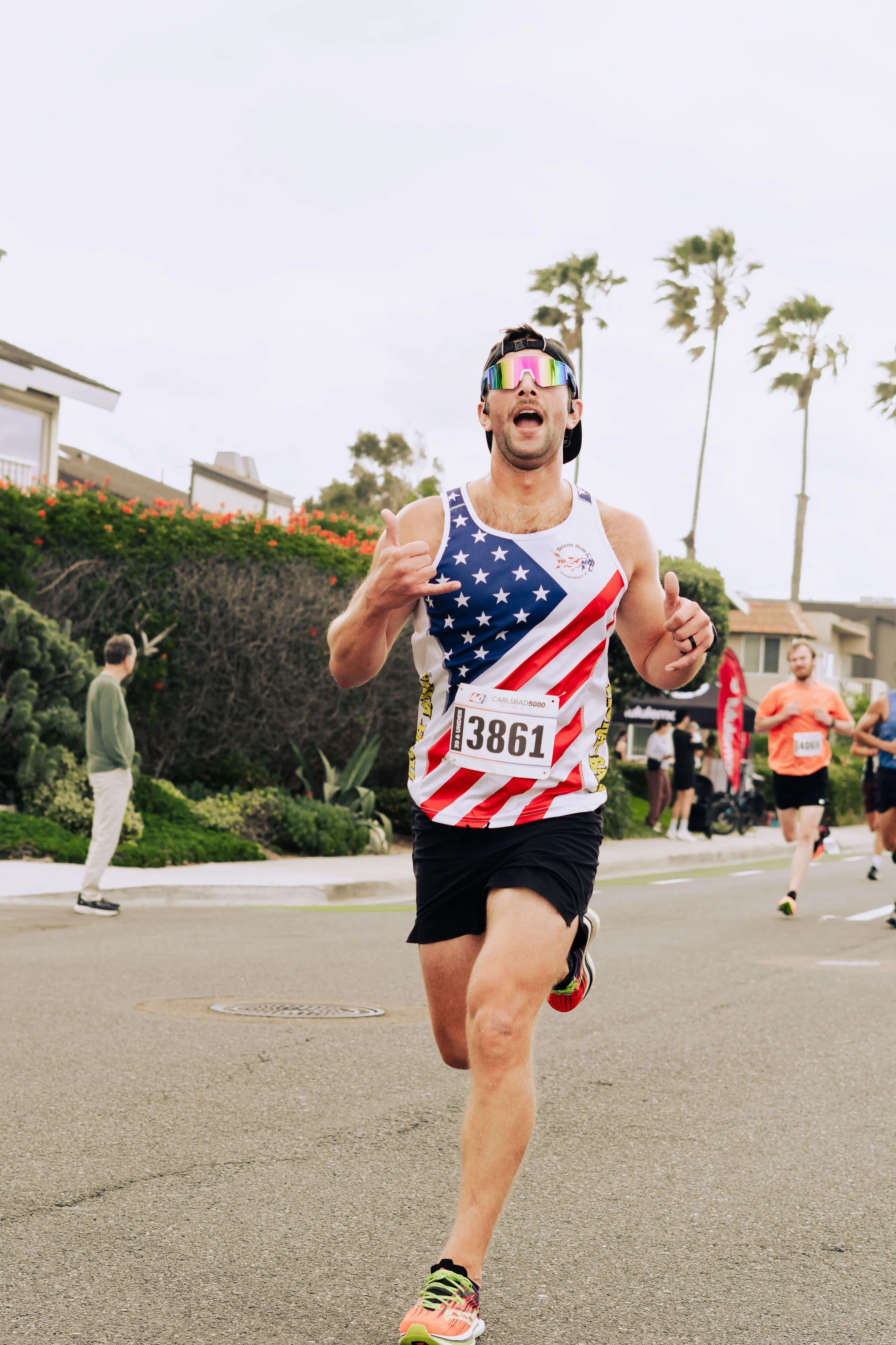 A man running in a race, wearing sunglasses with a reflective rainbow surface, a tank top with an American flag design, bib number 3861, and colorful running shoes, with houses and palm trees in the background.