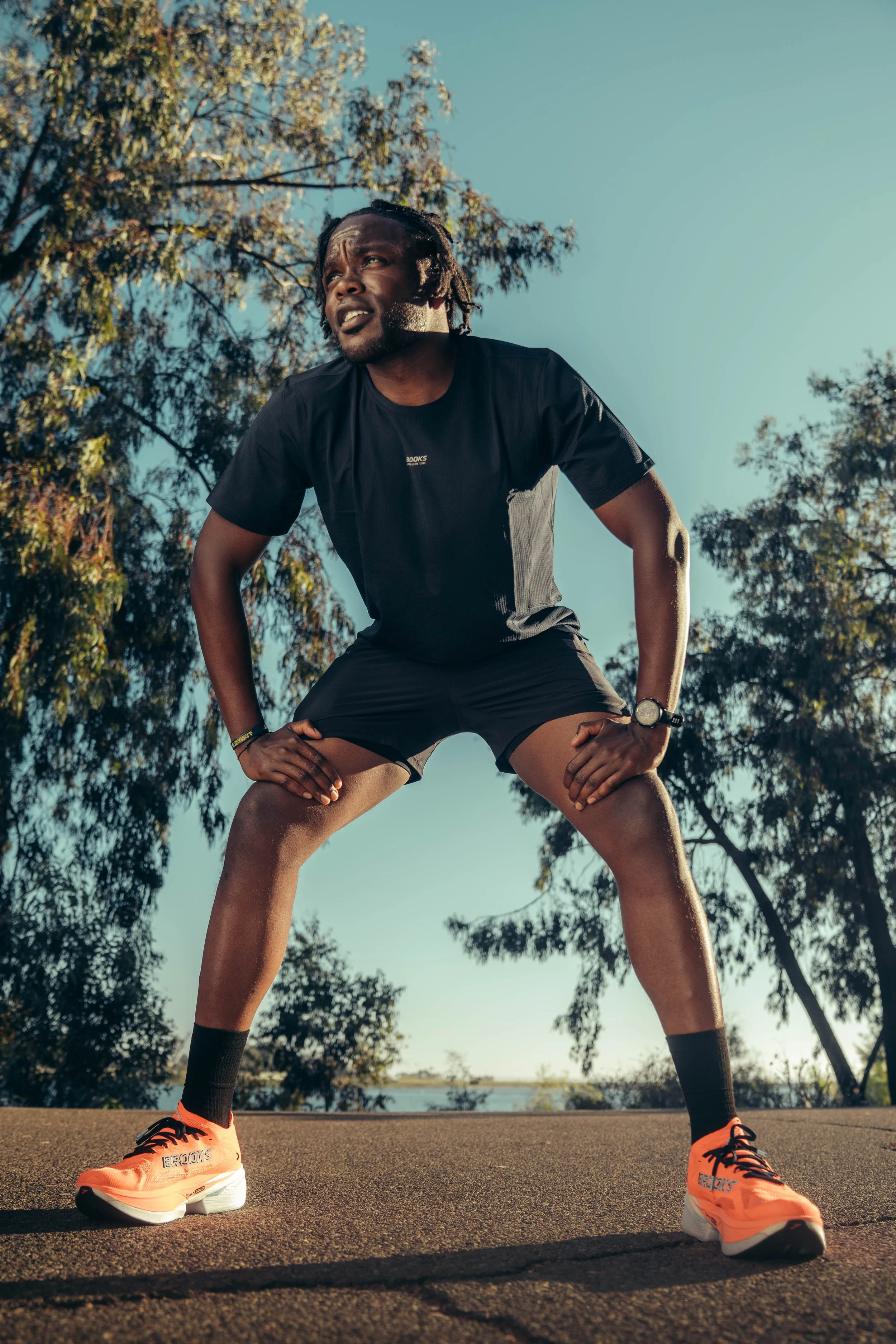 A man in athletic attire, including orange running shoes, black shorts, and a black shirt, is in a squat position outdoors with hands on knees, looking upward with trees and a body of water in the background.
