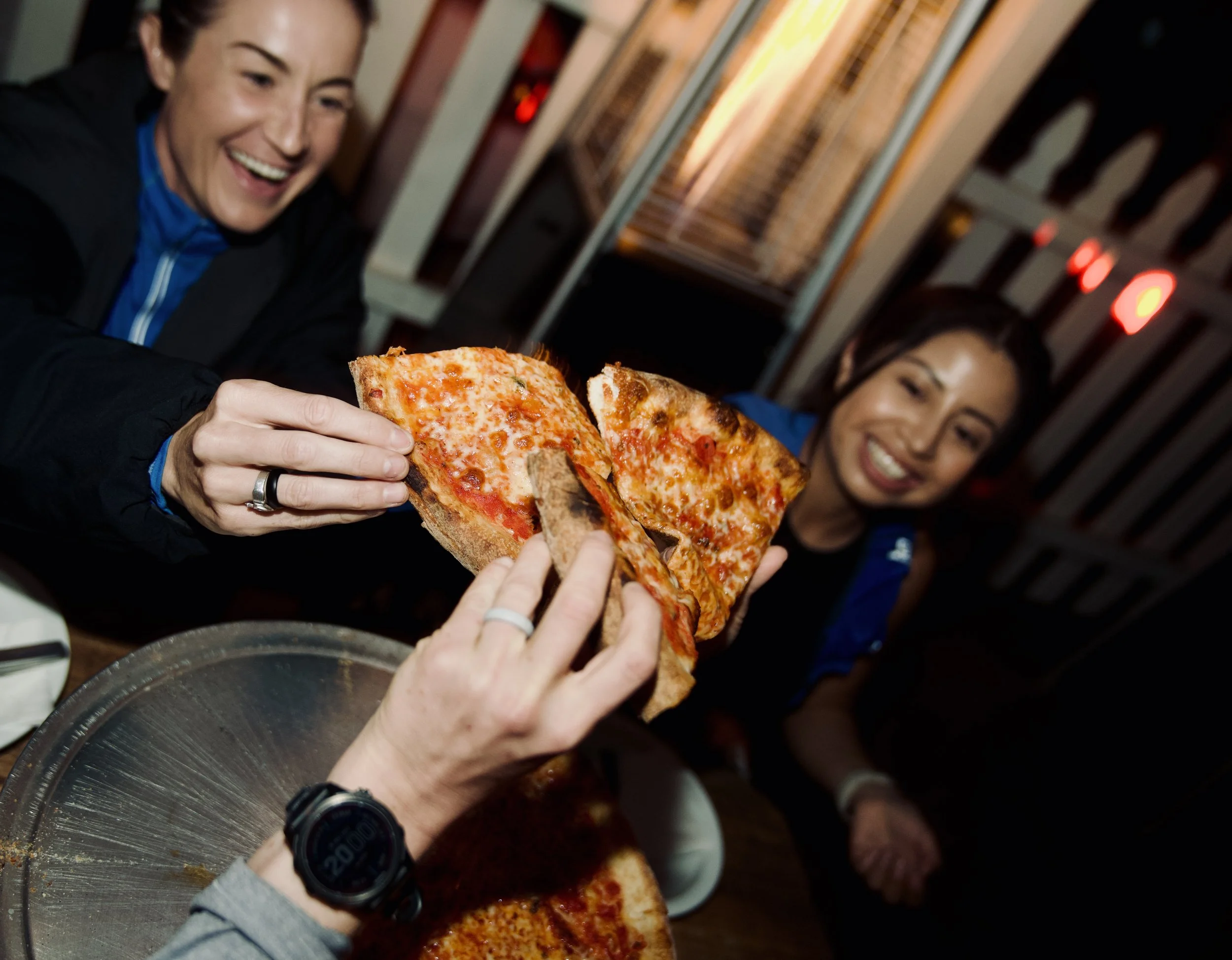 Two smiling people sharing slices of pepperoni pizza in a restaurant, with a person wearing a watch reaching for the pizza.