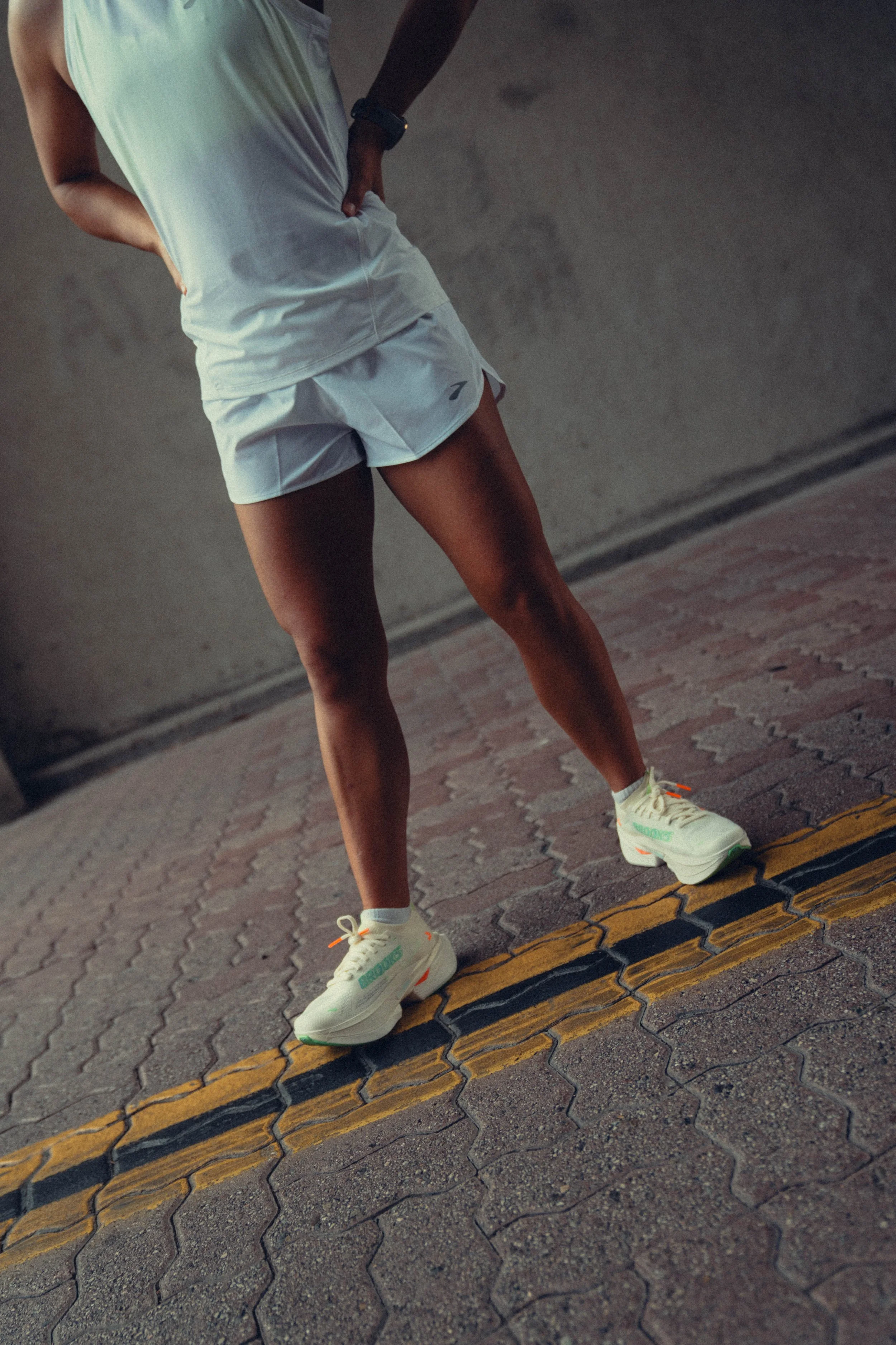 A person standing on a paved surface wearing white athletic shorts, a white shirt, and white sneakers, with their hands on their hips and one leg slightly bent.