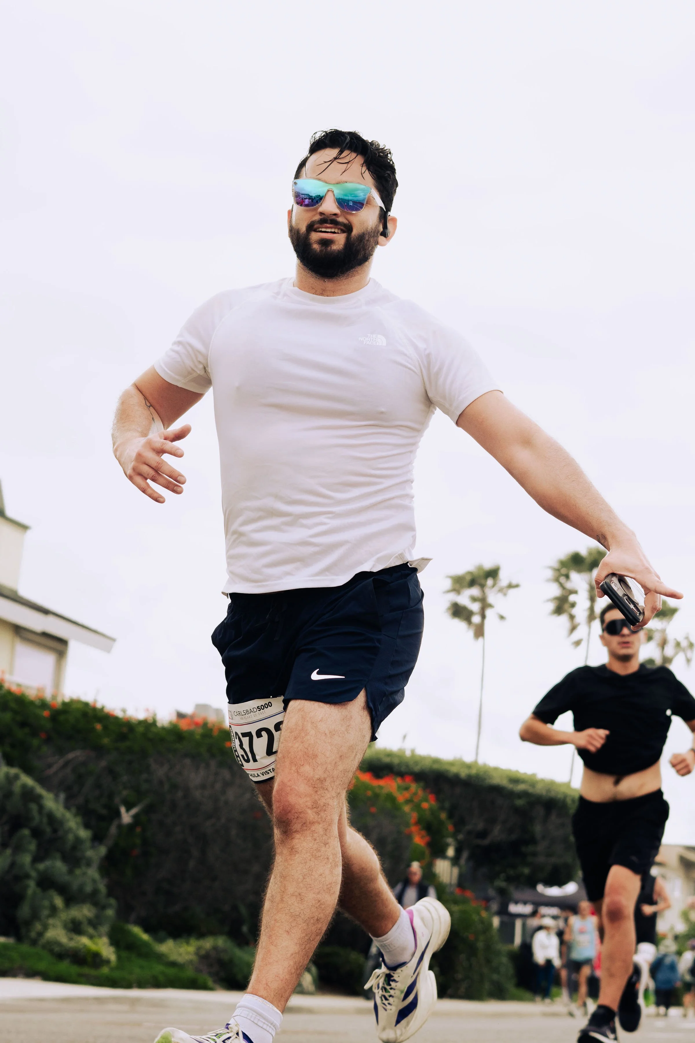 Man running outdoors wearing white t-shirt, navy shorts, sunglasses, and a race bib, with others running in the background on a cloudy day.