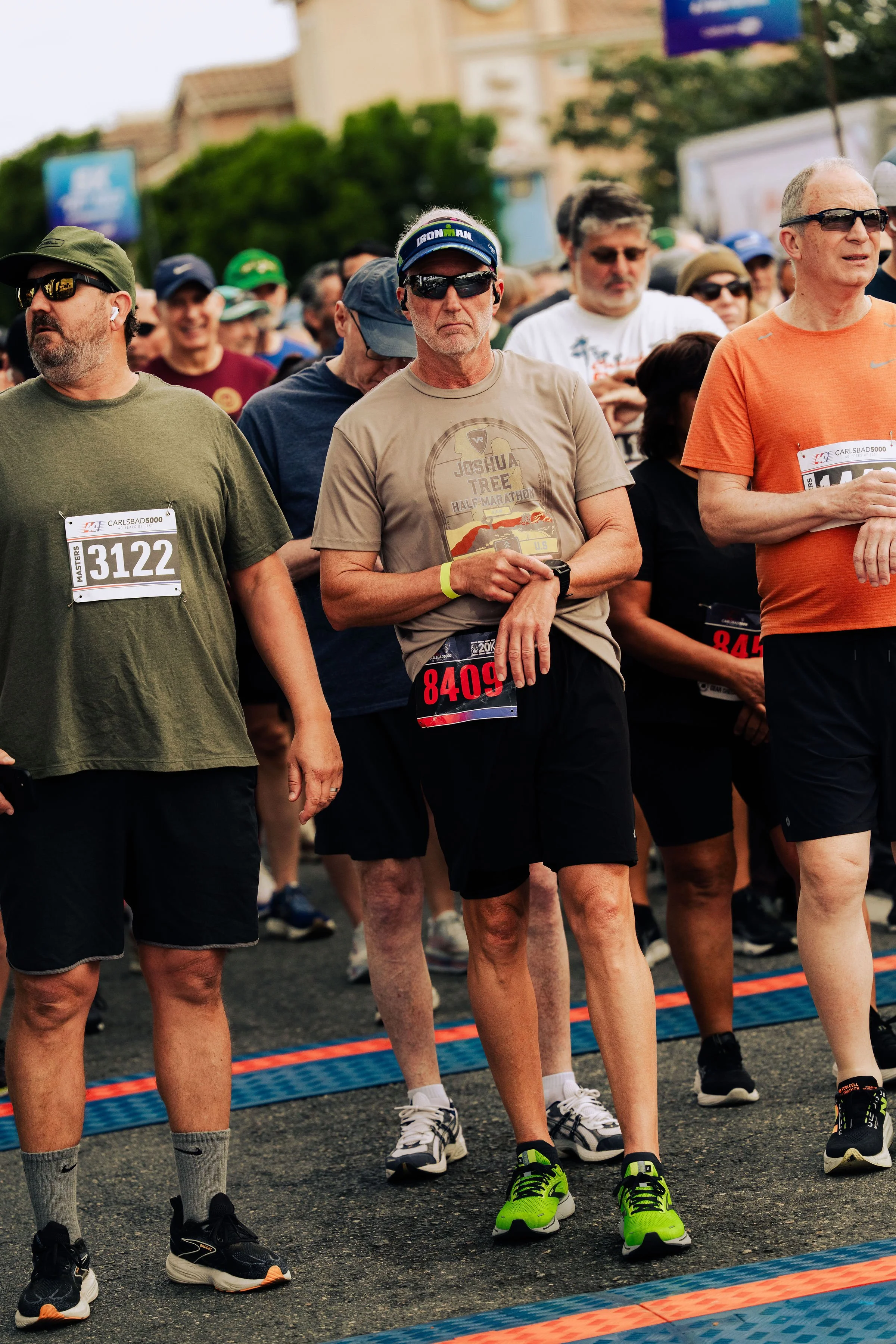 Crowd of runners waiting at the start line for a race, wearing athletic clothing and race bibs, with trees and buildings in the background.