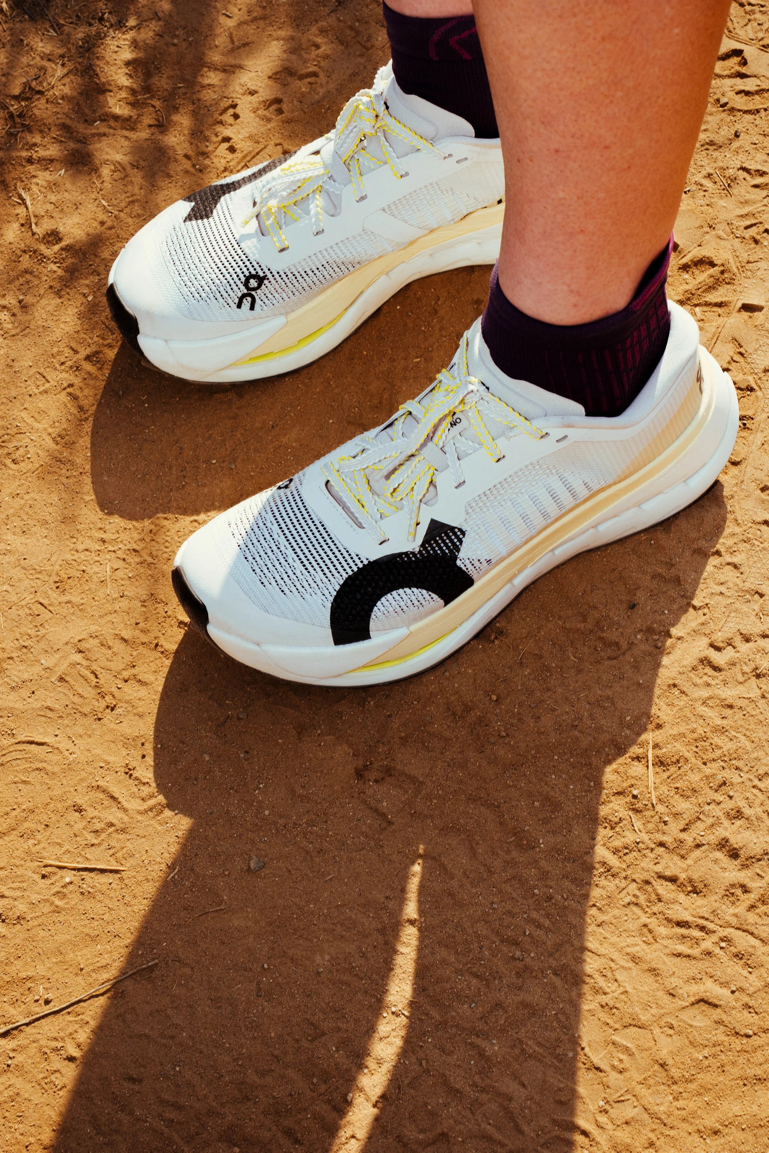 Person wearing white athletic shoes with yellow laces standing on sandy ground, casting a shadow.
